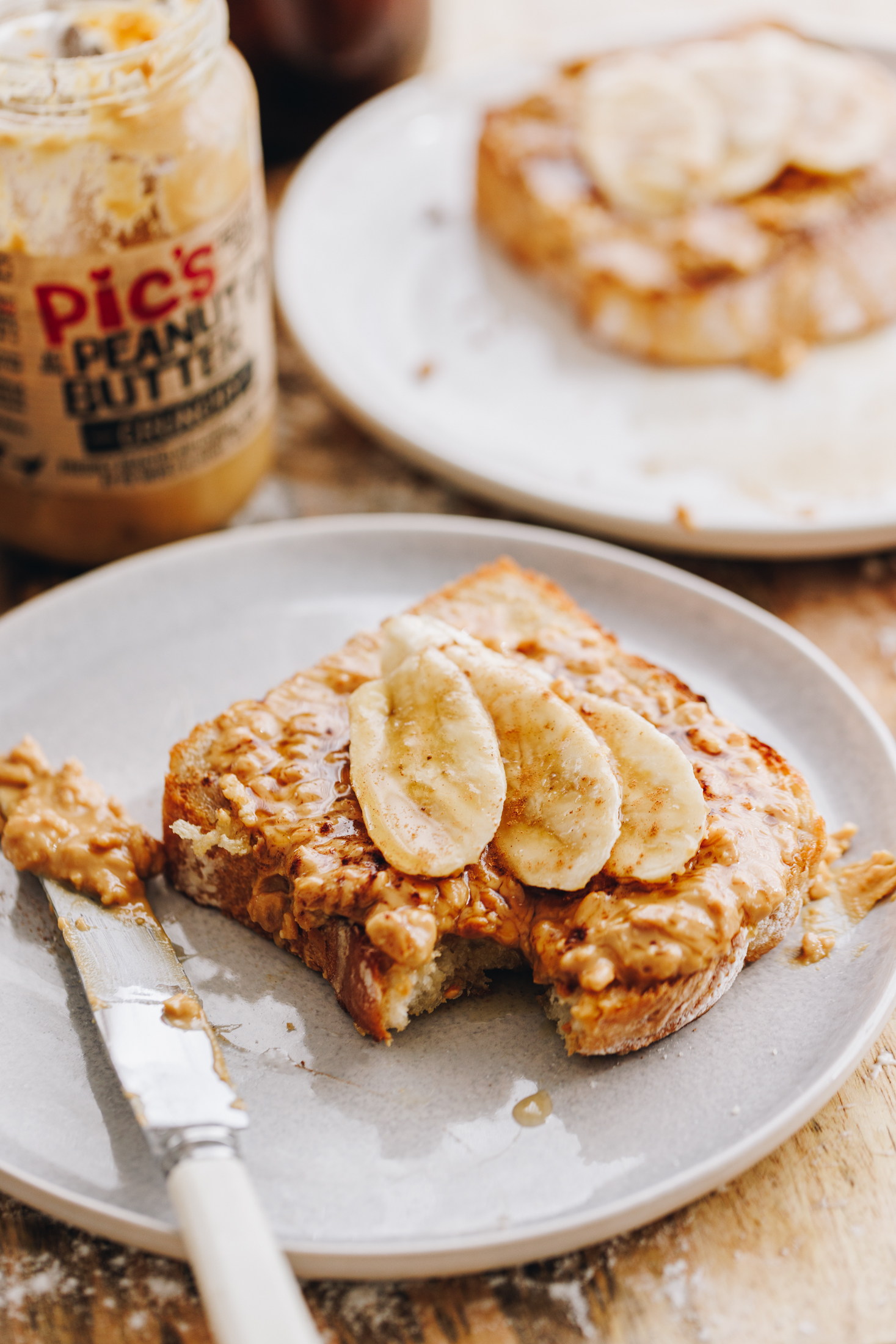 A peanut butter and banana toast using the no-knead white bread loaf is on a white plate. A jar of Pic's peanut butter is in the background. The toast has a bite out of it. 
