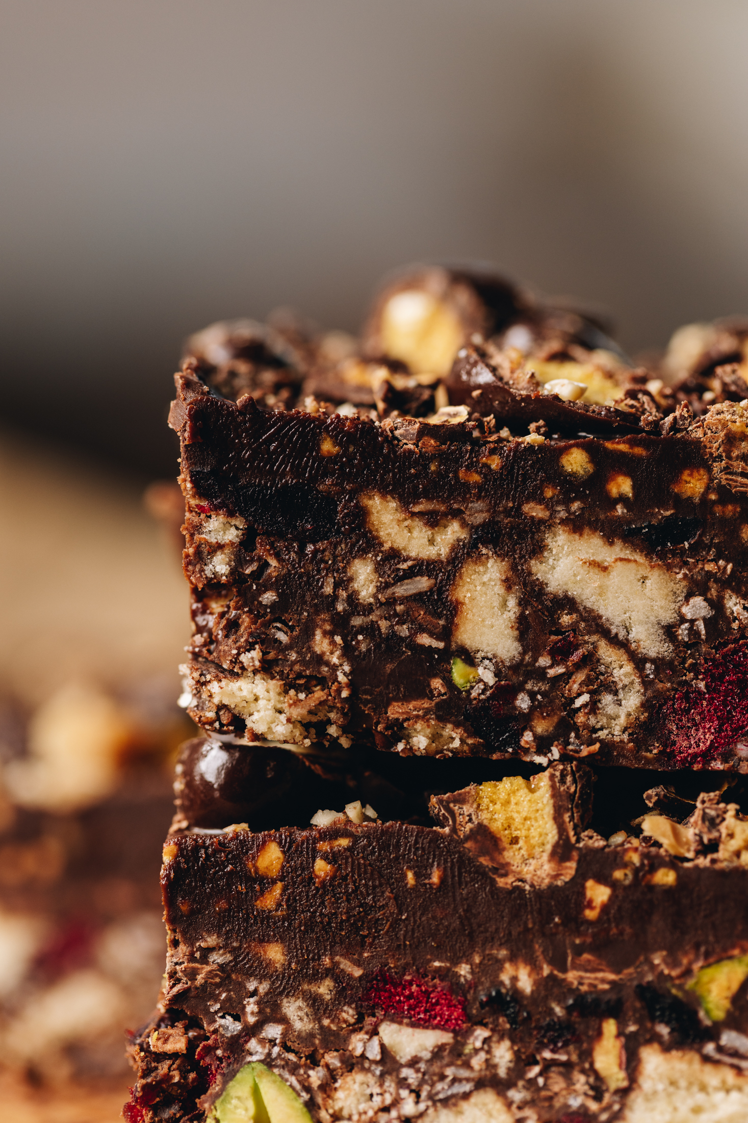 A close up shows two piece of no-bake hedgehog slice that is piled on top of each other. The slices have piece of chopped biscuit, freeze-dried fruit, pieces of pistachios and a chocolate topping with crushed chocolate peanuts on top. 
