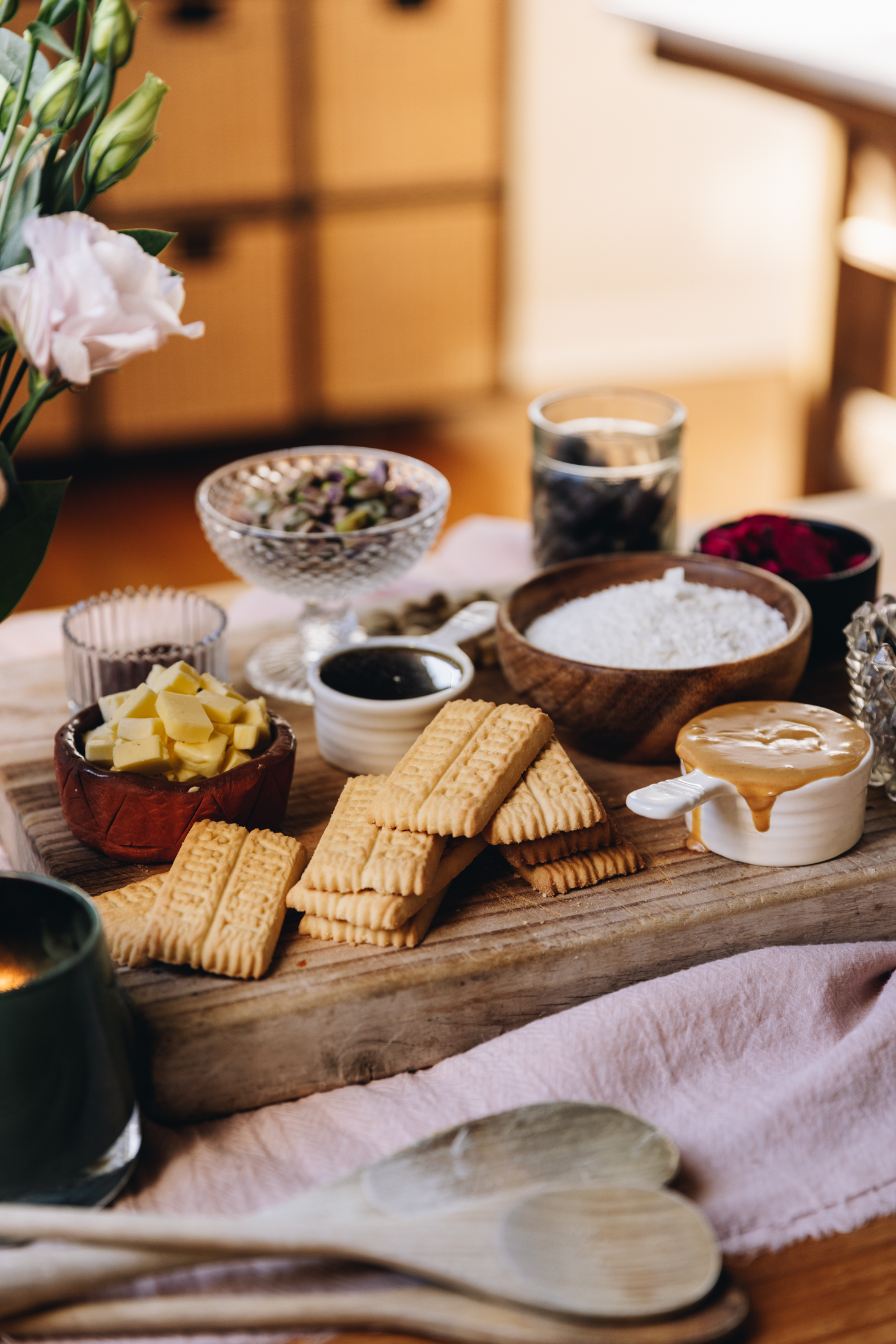 On a wooden table is a wooden board that is sitting on a pink cloth. Wooden spoons sit in front of the board and pink flowers are in a glass jar. On the board is scotch biscuits and different vintage bowls and cups. There is peanut butter, butter, pistachios, coconut, peanut butter and freeze-dried plums in view.