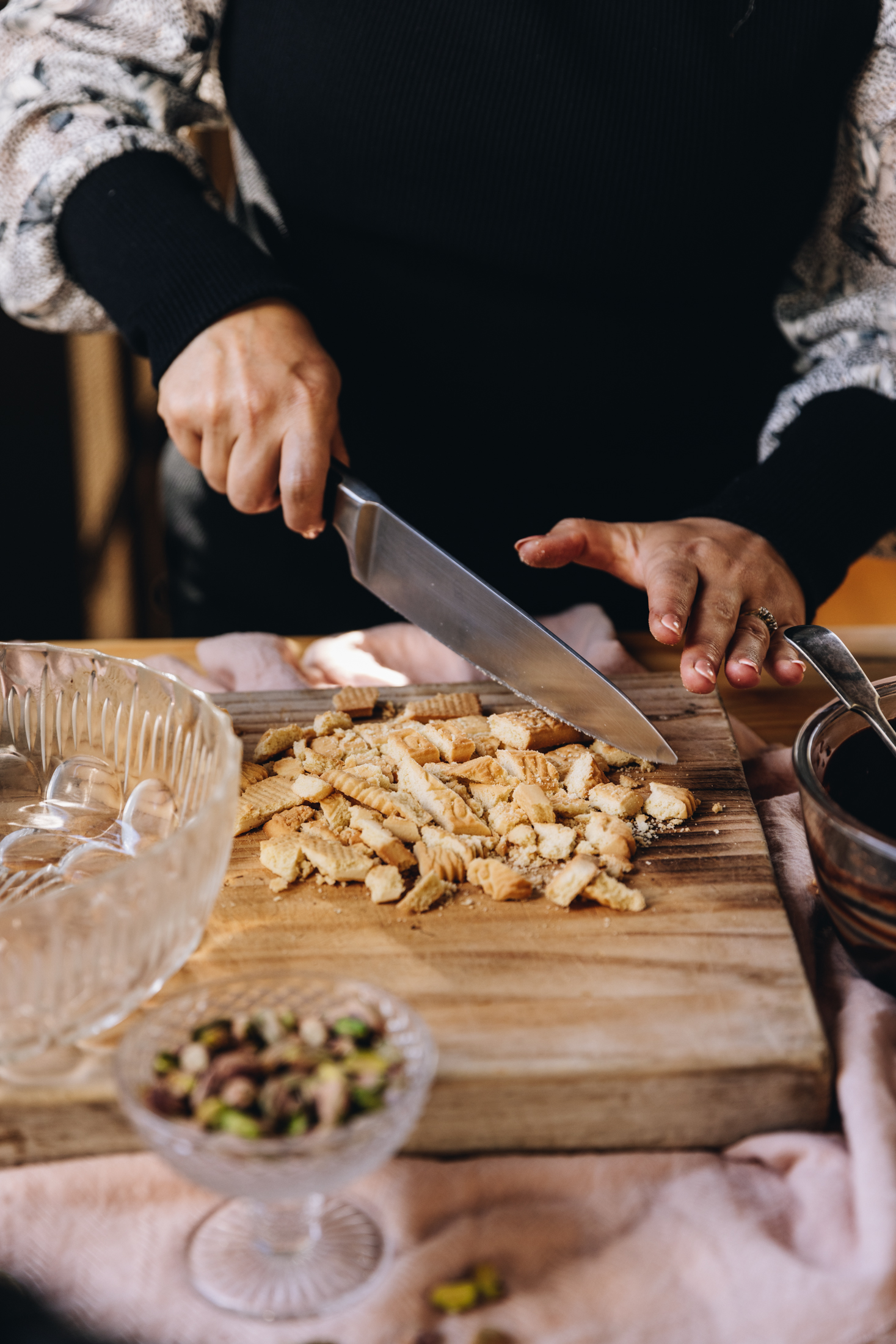 On a wooden table is a wooden board that is sitting on a pink cloth. Naomi is chopping Scott biscuits with a large knife on the board. A glass vintage bowl is also on the board. A small vintage glass bowl has pistachios in it, sits in front of the board.
