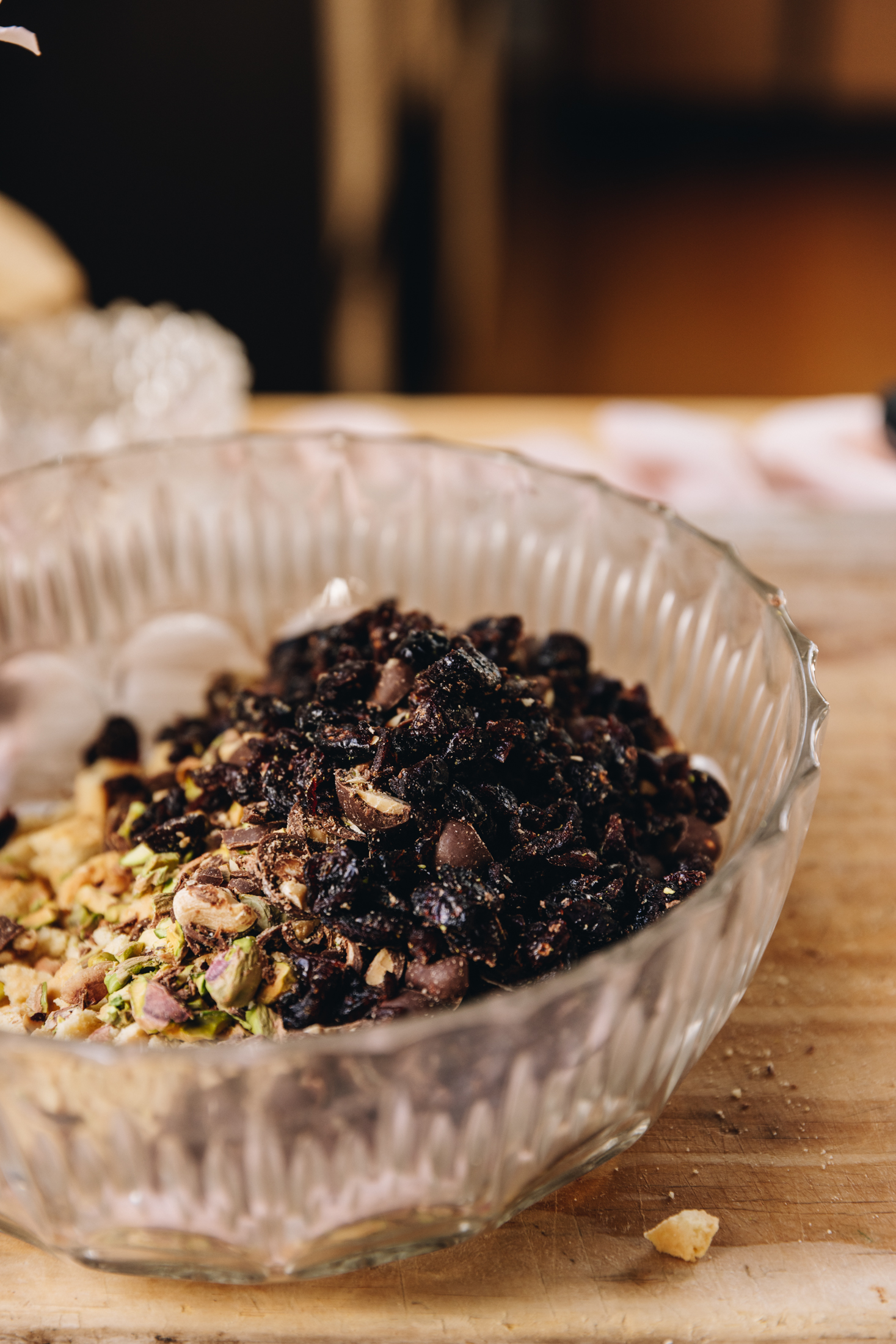 In a glass vintage bowl is hedgehog slice ingredients. Chopped cranberries, pistachios and biscuits can be sweet. The bowl is on a wooden table.