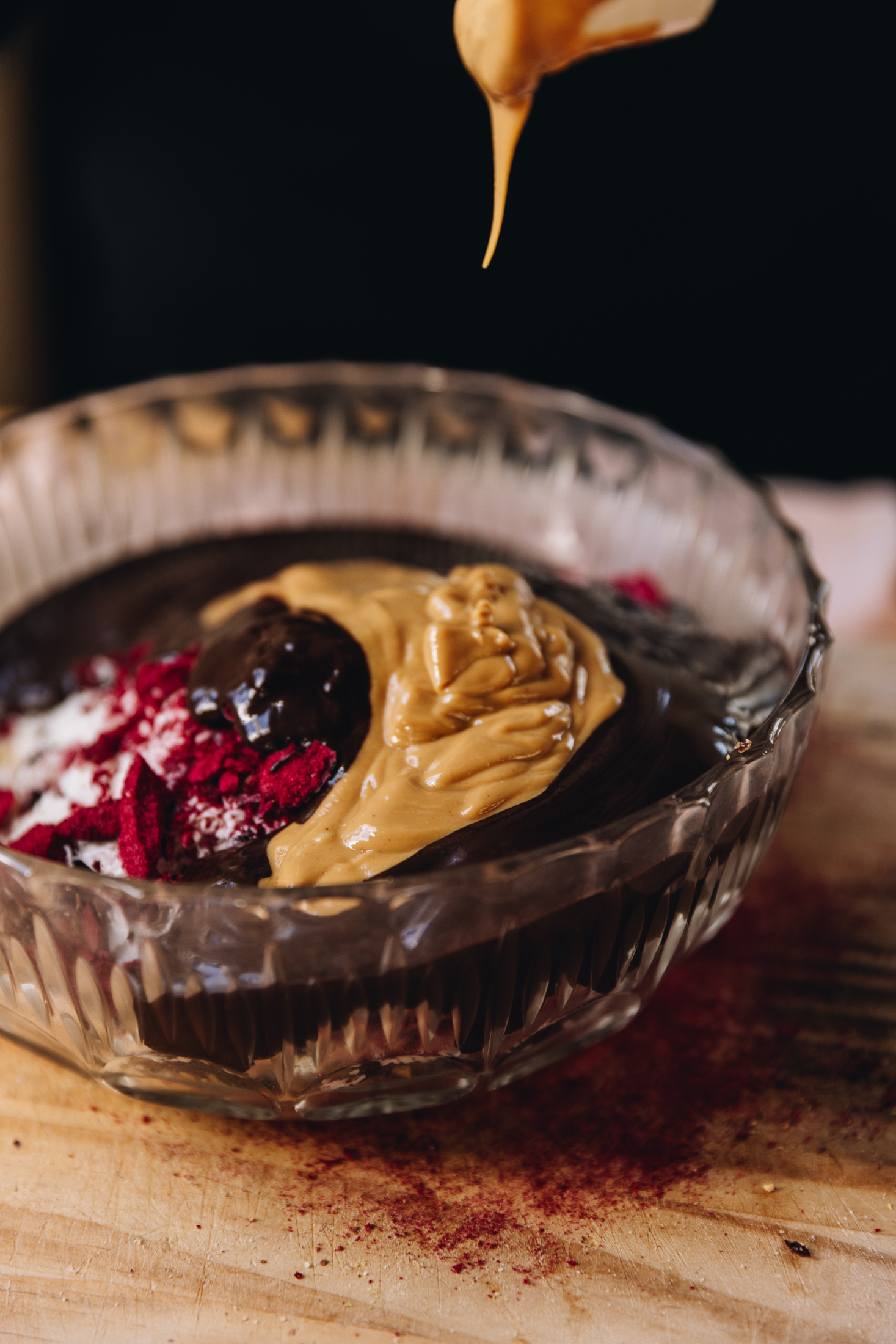 In a glass vintage bowl is hedgehog slice ingredients. Crushed freeze-dried plums, melted chocolate and runny smooth peanut butter are seen. It is on the wooden table.