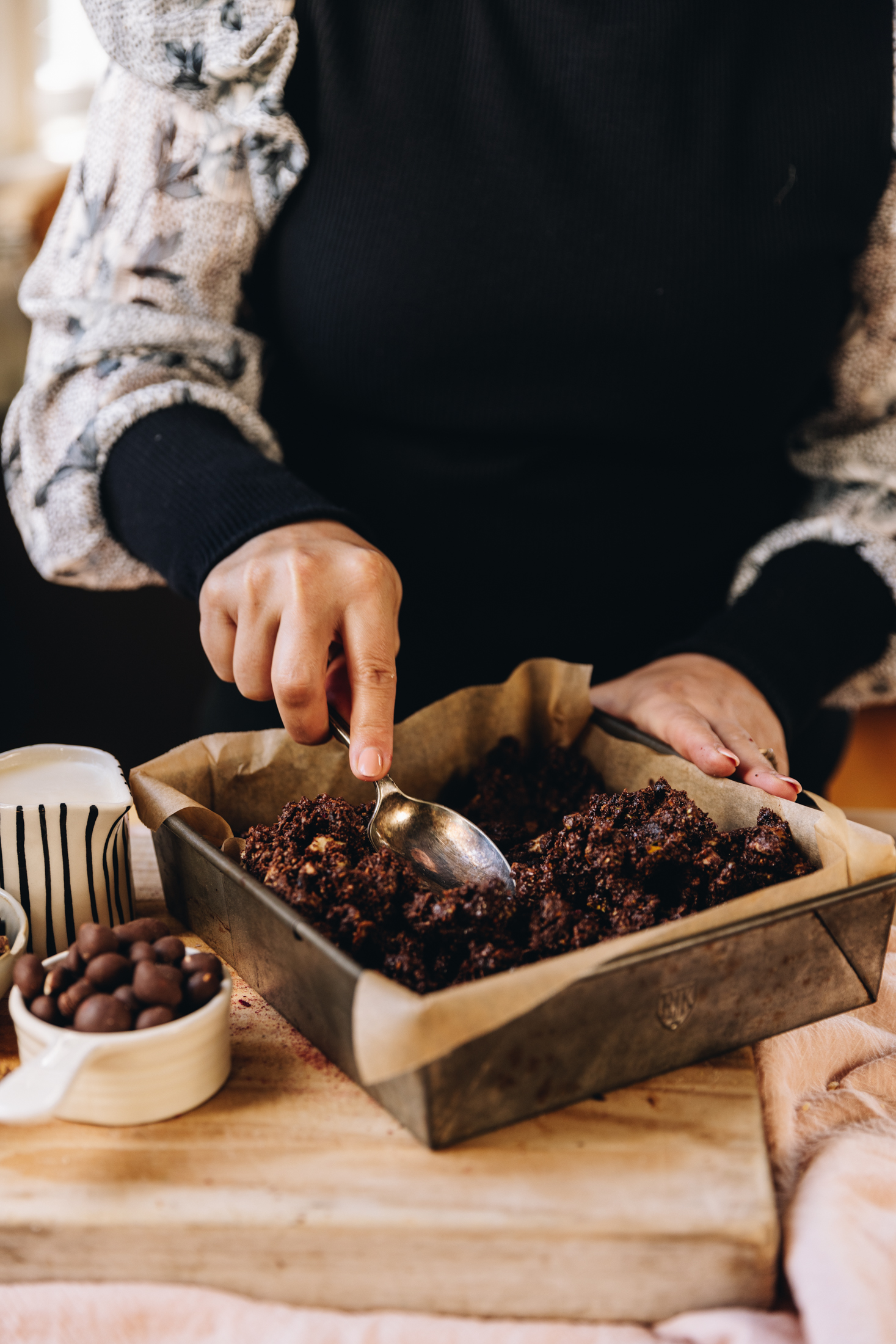 On a wooden table sits a vintage baking tin lined with baking paper. It has hedgehog slice filling in it and Naomi is pressing it down with a large spoon. Chocolate peanuts are in a cup next to it along with a small black and white striped jug.