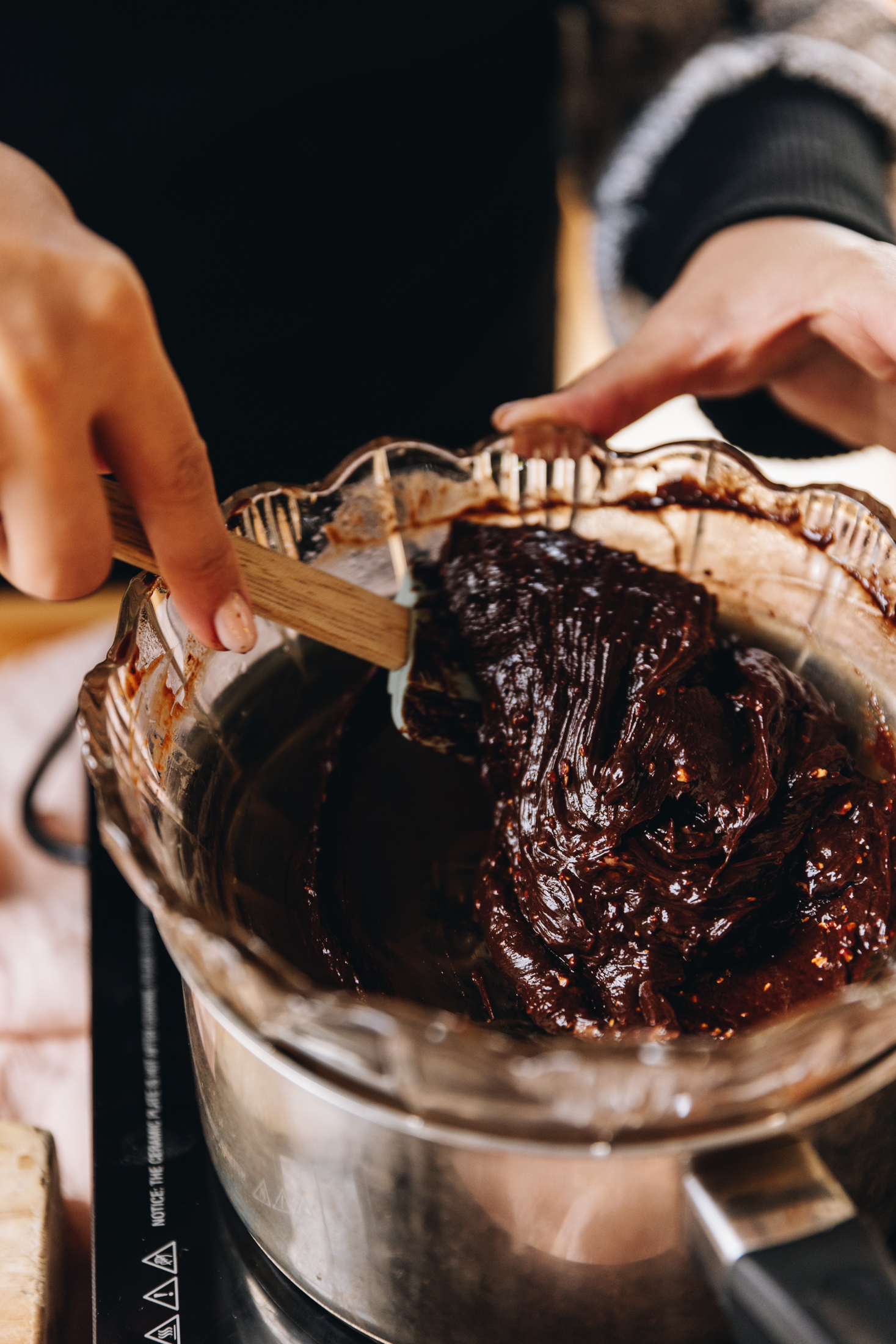 A silver pot sits on a black portable stove. It has a glass vintage bowl sitting in it with melted Hokey Pokey chocolate. Naomi is stirring it with a spatula.
