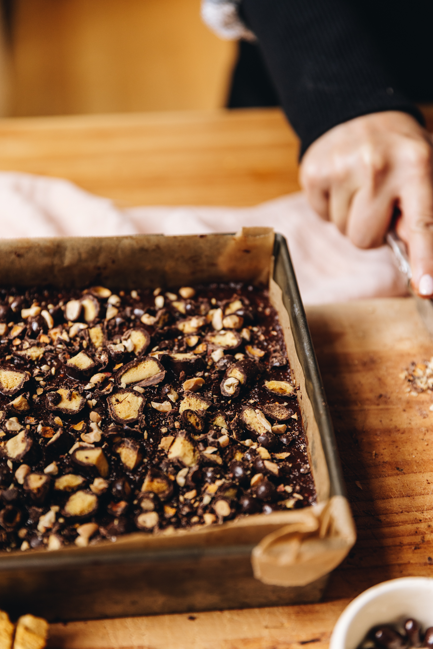 On a wooden table sits a vintage baking tin lined with baking paper. It has a no-bake hedgehog slice in it and is topped with chocolate and chocolate covered chopped peanuts and honeycomb. A pink cloth is in the background. 