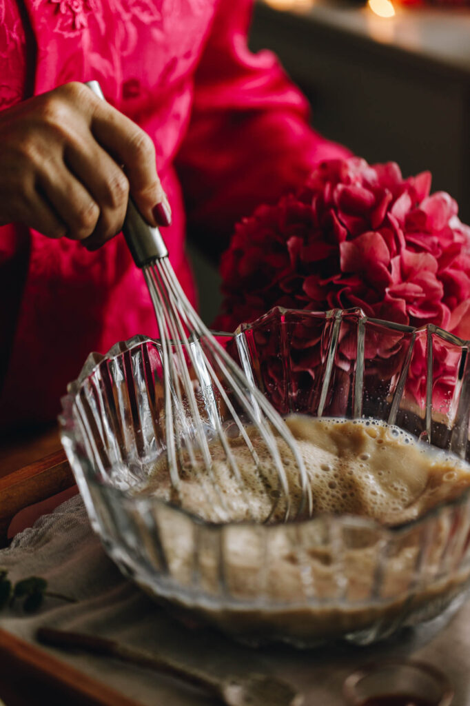 A glass bowl is filled with yeast mixture and is being stirred with a steel whisk. It sits on a wooden bench and there are pink flowers in the background.