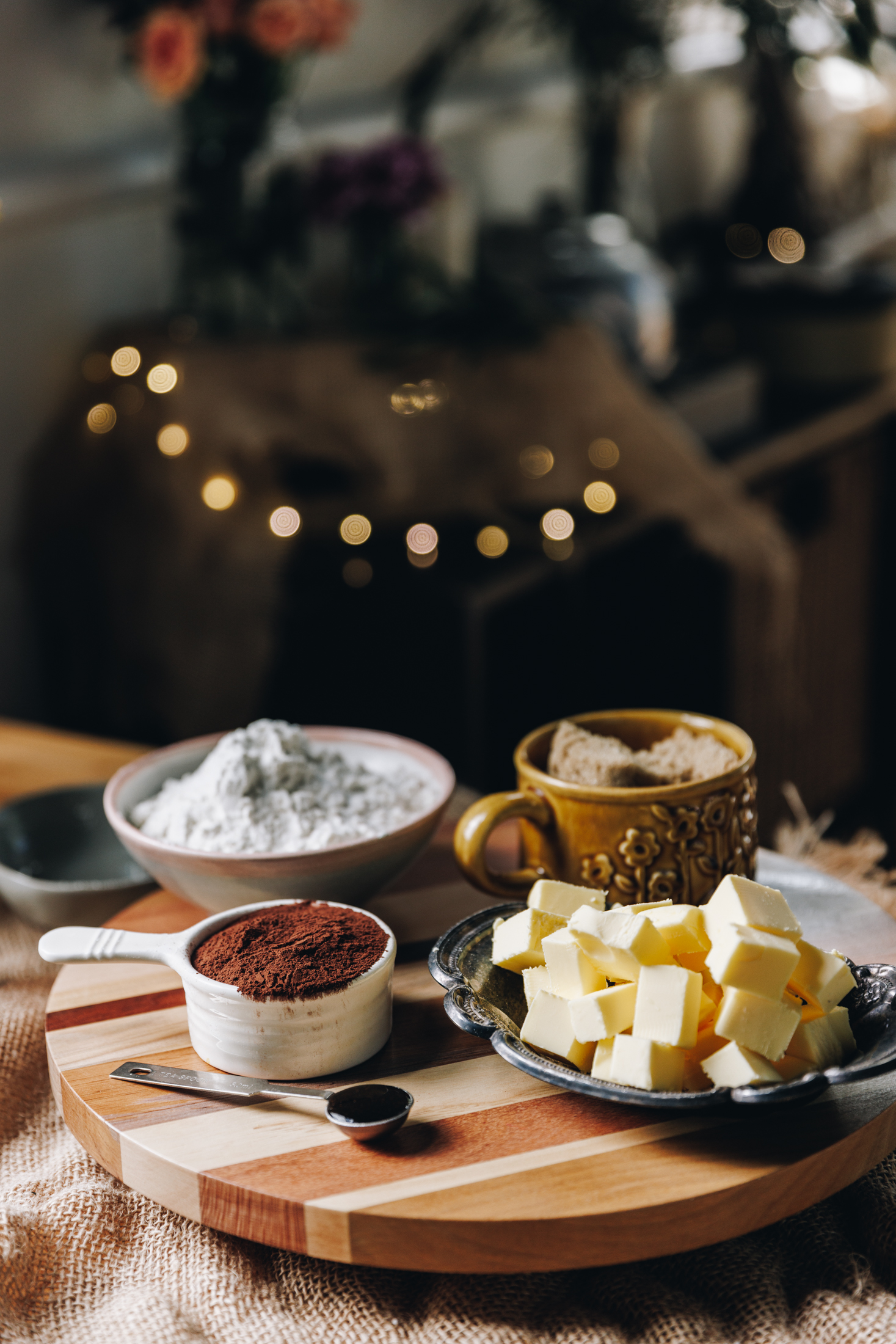 Cookie Ingredients are in different vintage bowls and are sitting on a wooden board on top of wooden table. There is fairy lights in the background.