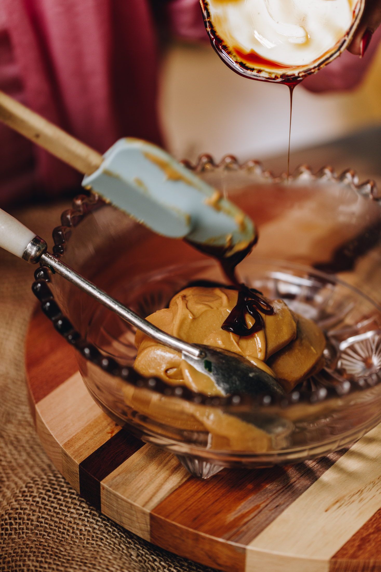 On a wooden board is a vintage bowl. In it is peanut butter and a spatula is adding date syrup. 