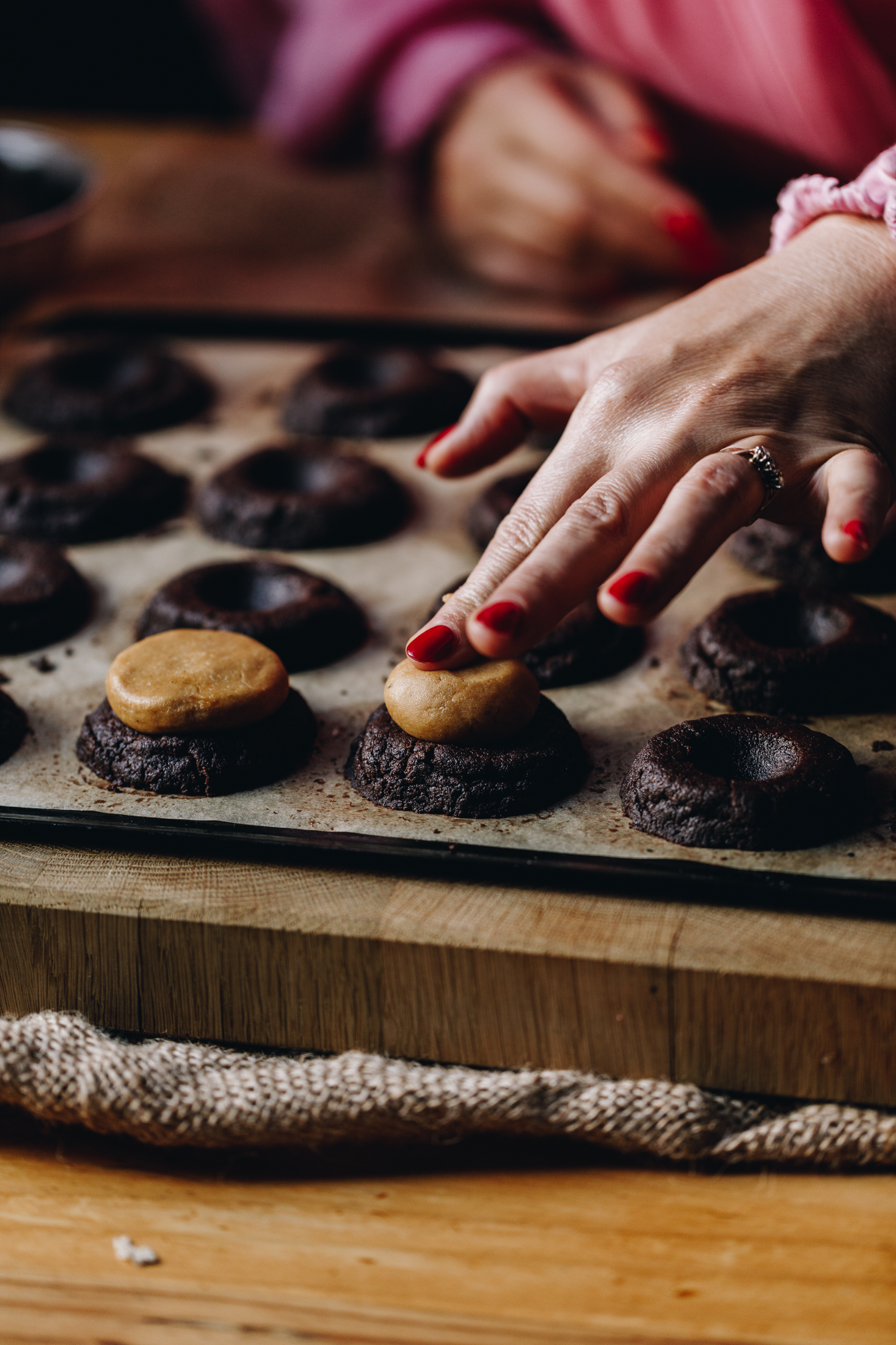 On a tray lined with baking paper is freshly baked Buckeye cookies. They have been indented with a teaspoon and a peanut butter truffle is being pressed in to one cookie. 