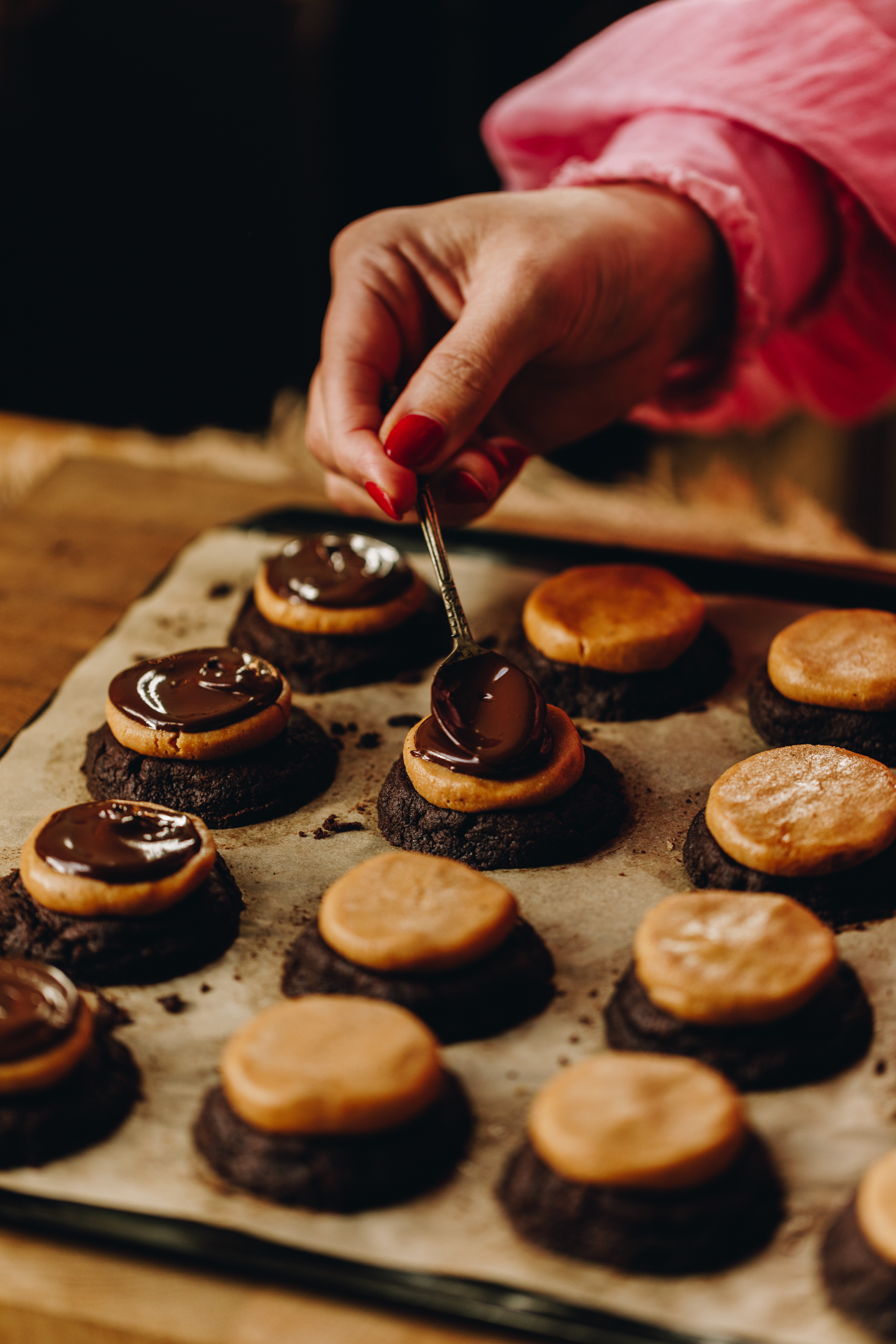 On a tray lined with baking paper is freshly baked Buckeye cookies. They have peanut butter truffles top and chocolate is spread on some. A teaspoon is being spread on to one and some are still plain.