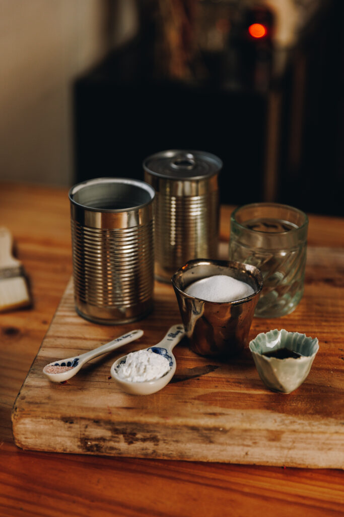 The Coconut Buns (Pani Popo) coconut sauce ingredients are added to a wooden board, sitting on a wooden table. Each ingredient is added in to different antique vessels and cans with the paper covering removed.