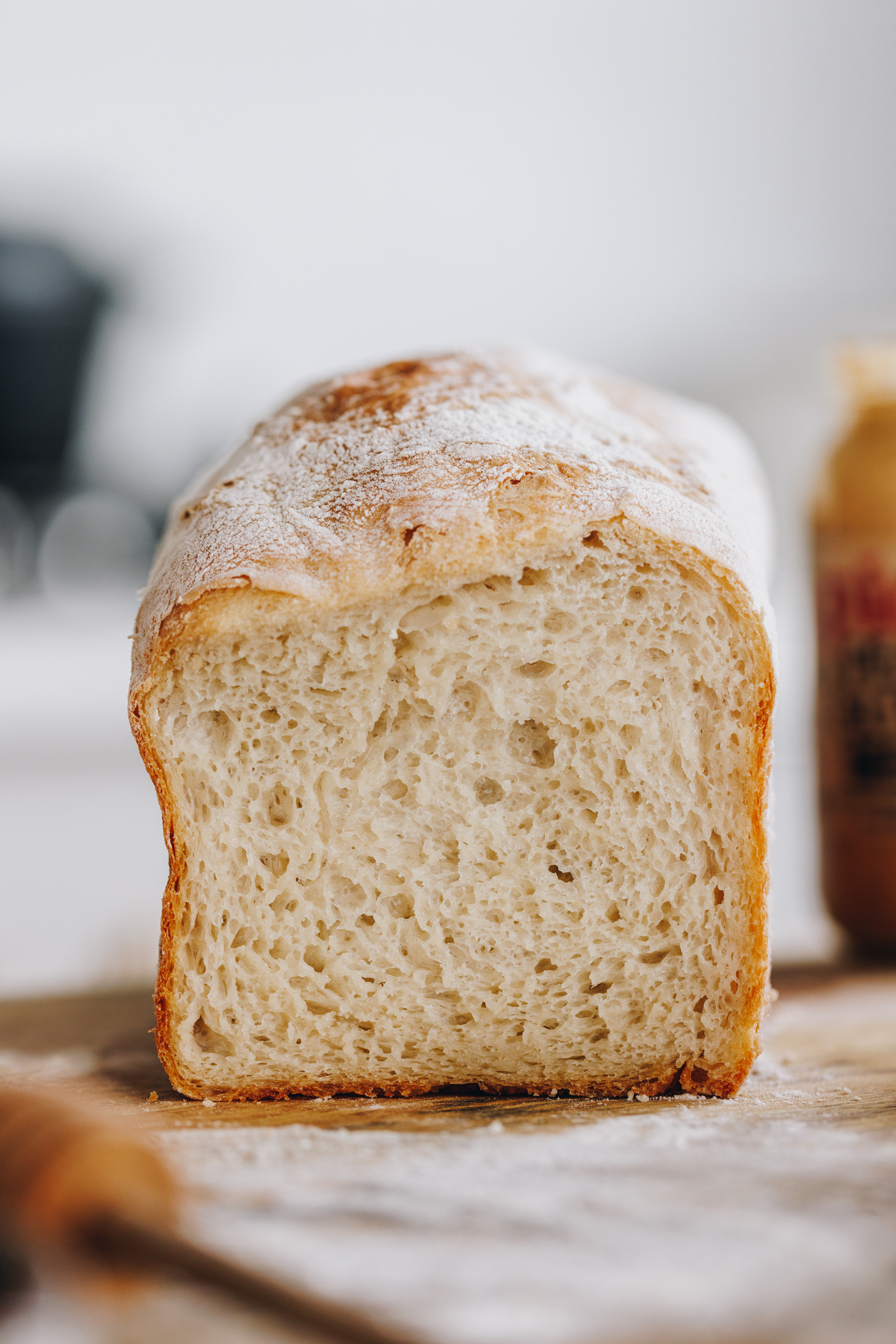 The No-knead bread loaf is on a wooden board that is dusted with flour. It is cut open revealing the soft, fluffy interior.
