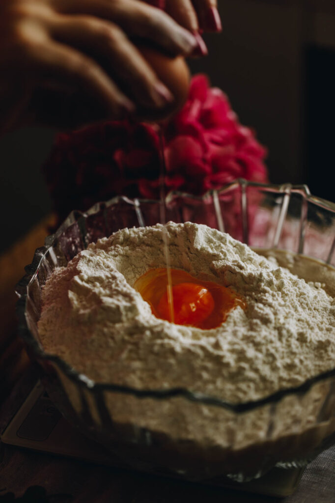 In to a glass bowl - an egg is being broken in to a large amount of flour. A pink flower is in the background.