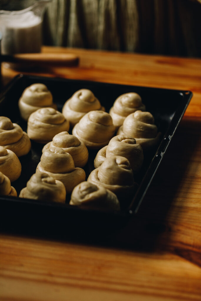 The unbaked Coconut Buns (Pani Popo) are rolled up and evenly spaced out in a black tray, sitting on a wooden table.