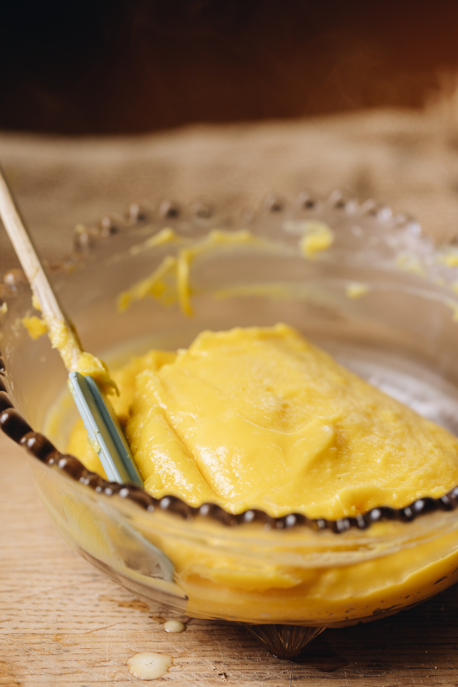 A simple lemon custard recipe is in a vintage purple bowl with a blue spatula with a wooden handle in it. It sits on a wooden table. 