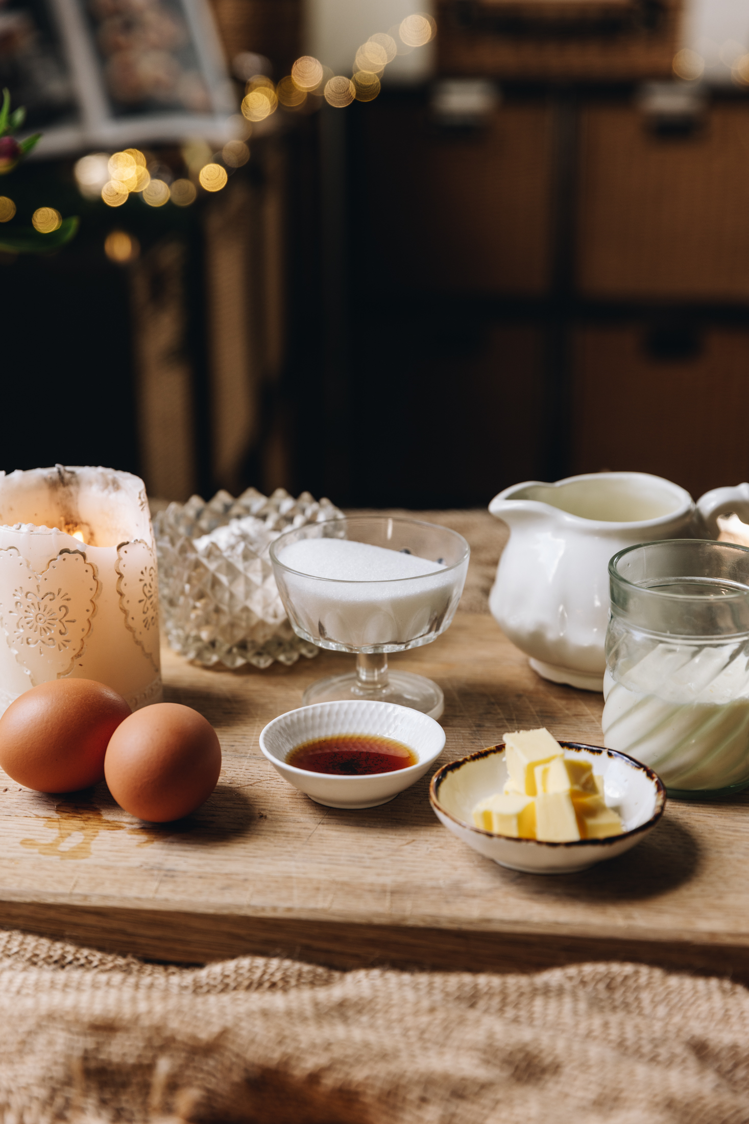 A wooden board sits on top of natural weaved fabric. On the board is whole eggs with butter, vanilla, milk, cream and sugar in vintage glasses, containers and jugs. A white candle is burning next to the eggs with fairy lights blurred in the background. 