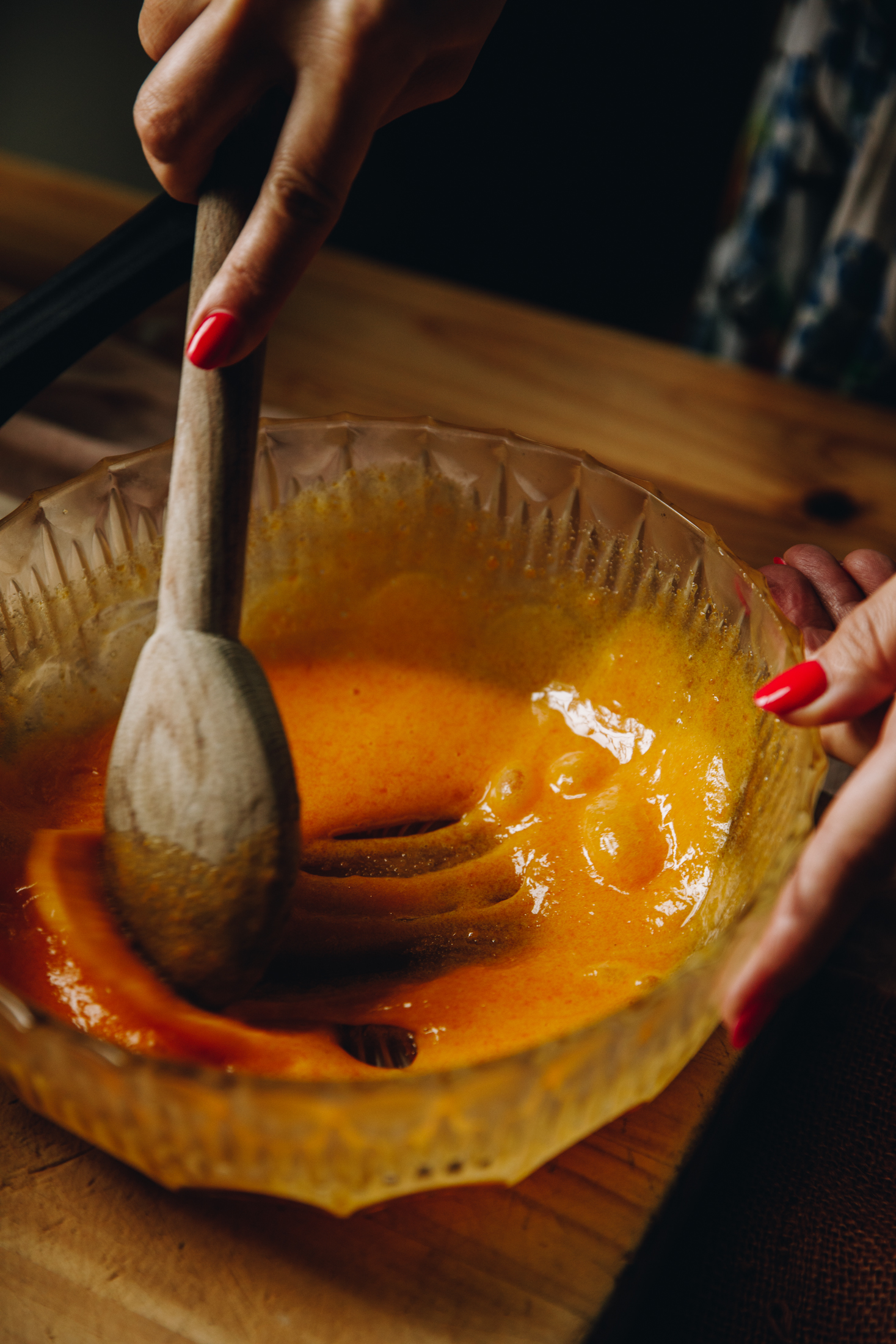 A glass vintage bowl is on a wooden table. Eggs are being mixed with a wooden spoon. 