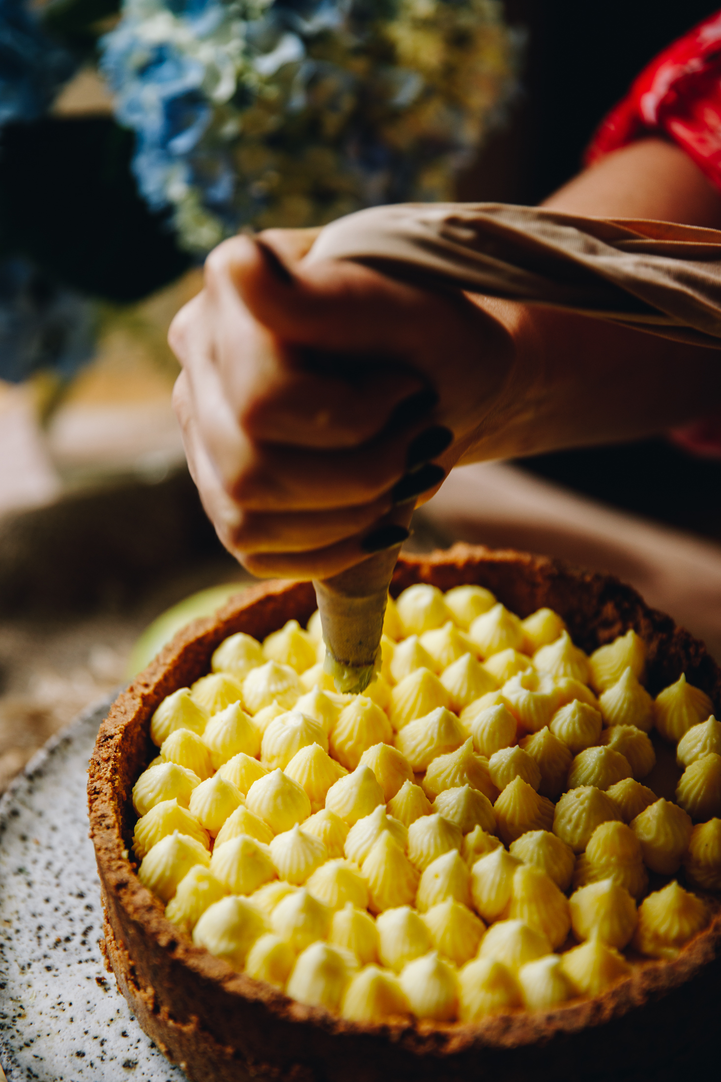 A cheesecake sits on a ceramic cake stand. Simple lemon custard is being piped in to the cheesecake. Blue flowers are in the background. 