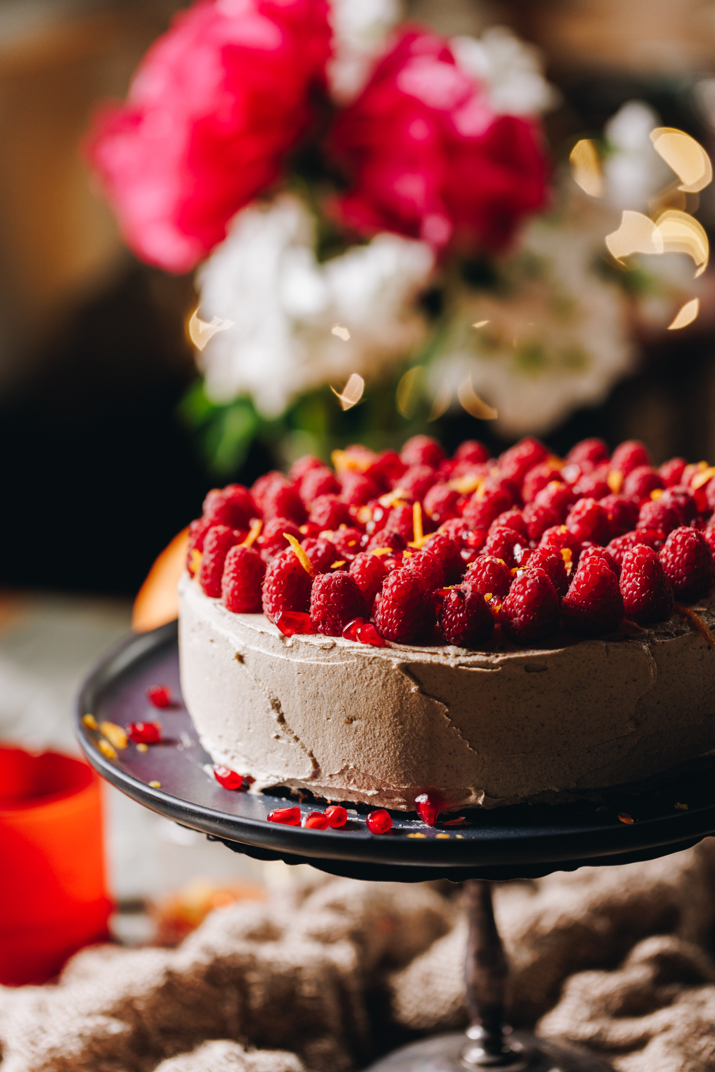A silver cake stand sits on a wooden table. On the cake stand is a whole orange cake that is decorated in cinnamon buttercream with fresh raspberries covering the top of the cake with grated orange zest. A large bunch of flowers are in the background.