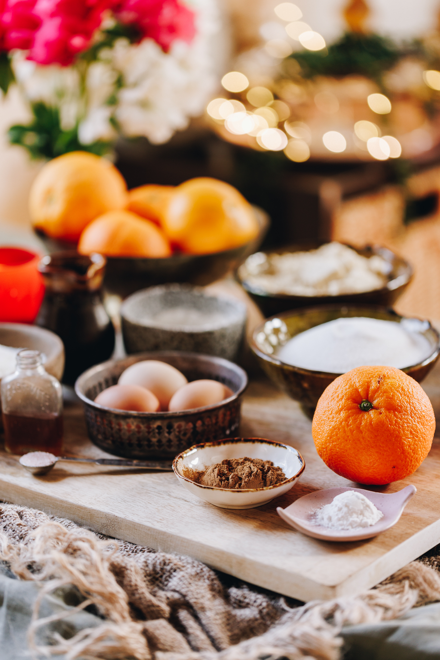 On a wooden board sits vintage bowls and small plates that have orange cake ingredients on it. There is oranges, eggs, spices and flour in view.
