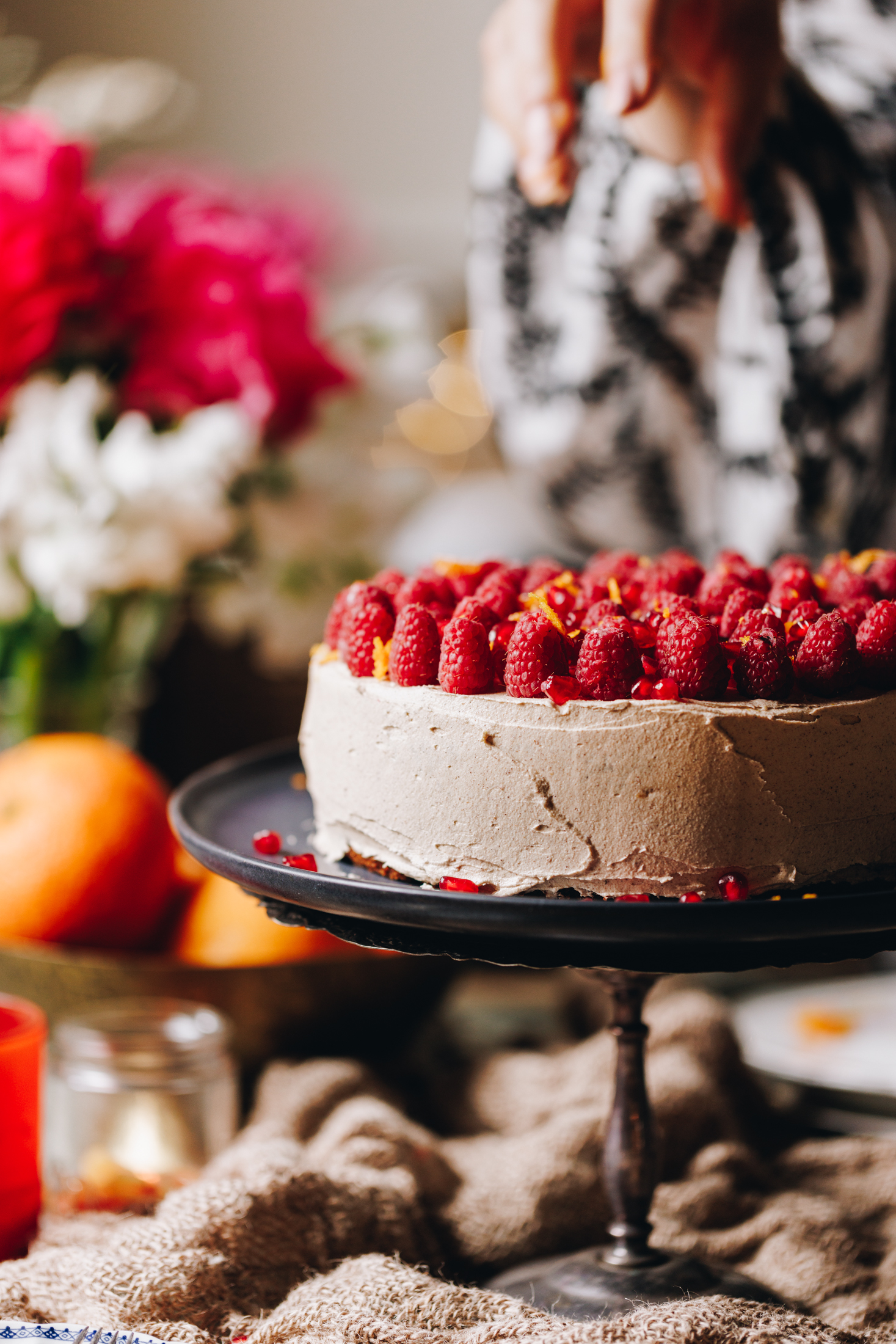 A silver cake stand sits on a wooden table. On the cake stand is a whole orange cake that is decorated in cinnamon buttercream with fresh raspberries covering the top of the cake with grated orange zest. A large bunch of flowers are in the background. A hand is sprinkling pomegranate seeds on to it. 