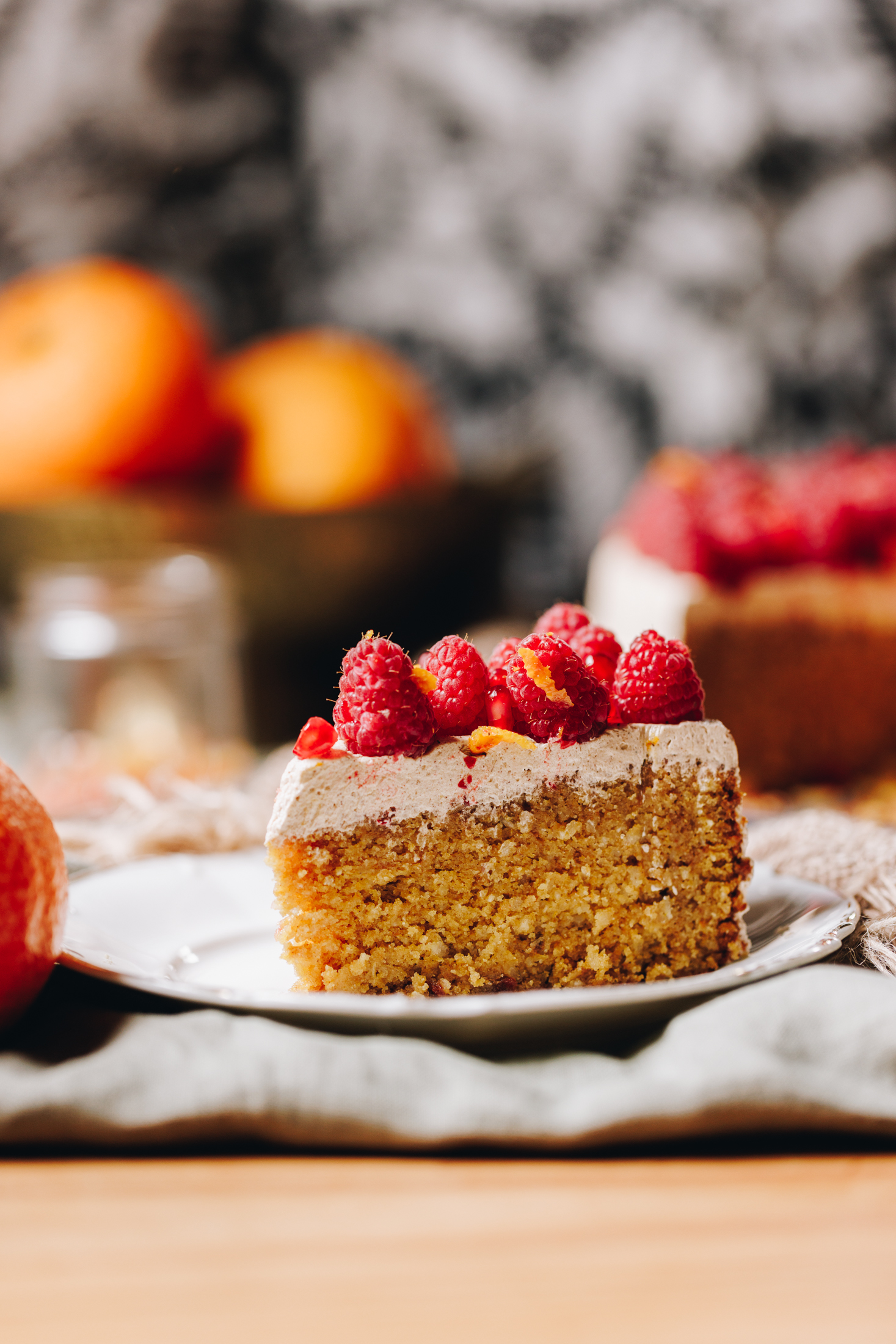 A slice of Whole Orange Almond Cake sits on a white small plate. It is on a wooden table on a soft green table cloth. The cake has cinnamon buttercream and fresh raspberries on top. 