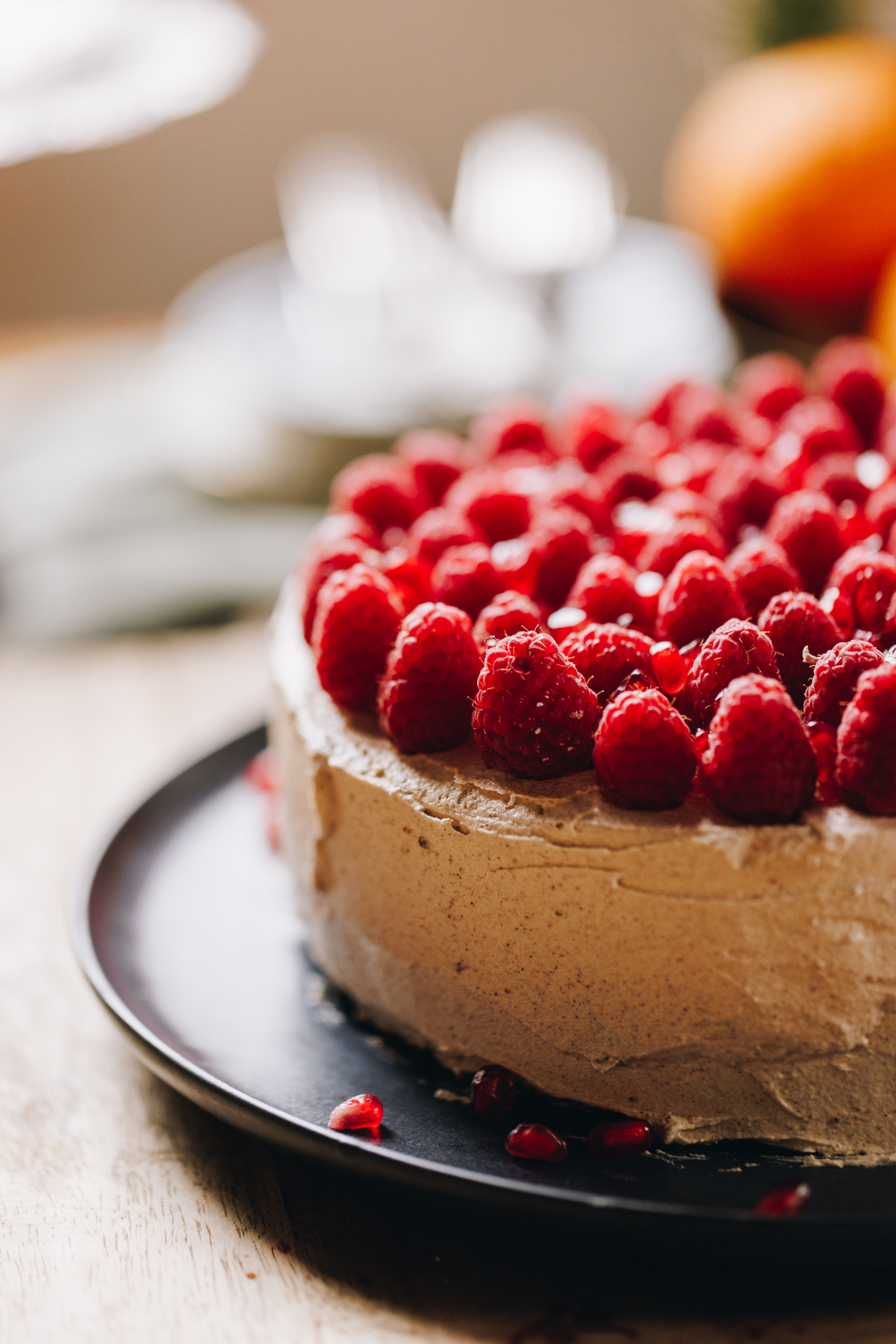 A blue plate sits on a wooden table. On the plate is a whole orange cake that is decorated in simple cinnamon buttercream with fresh raspberries covering the top of the cake with grated orange zest.