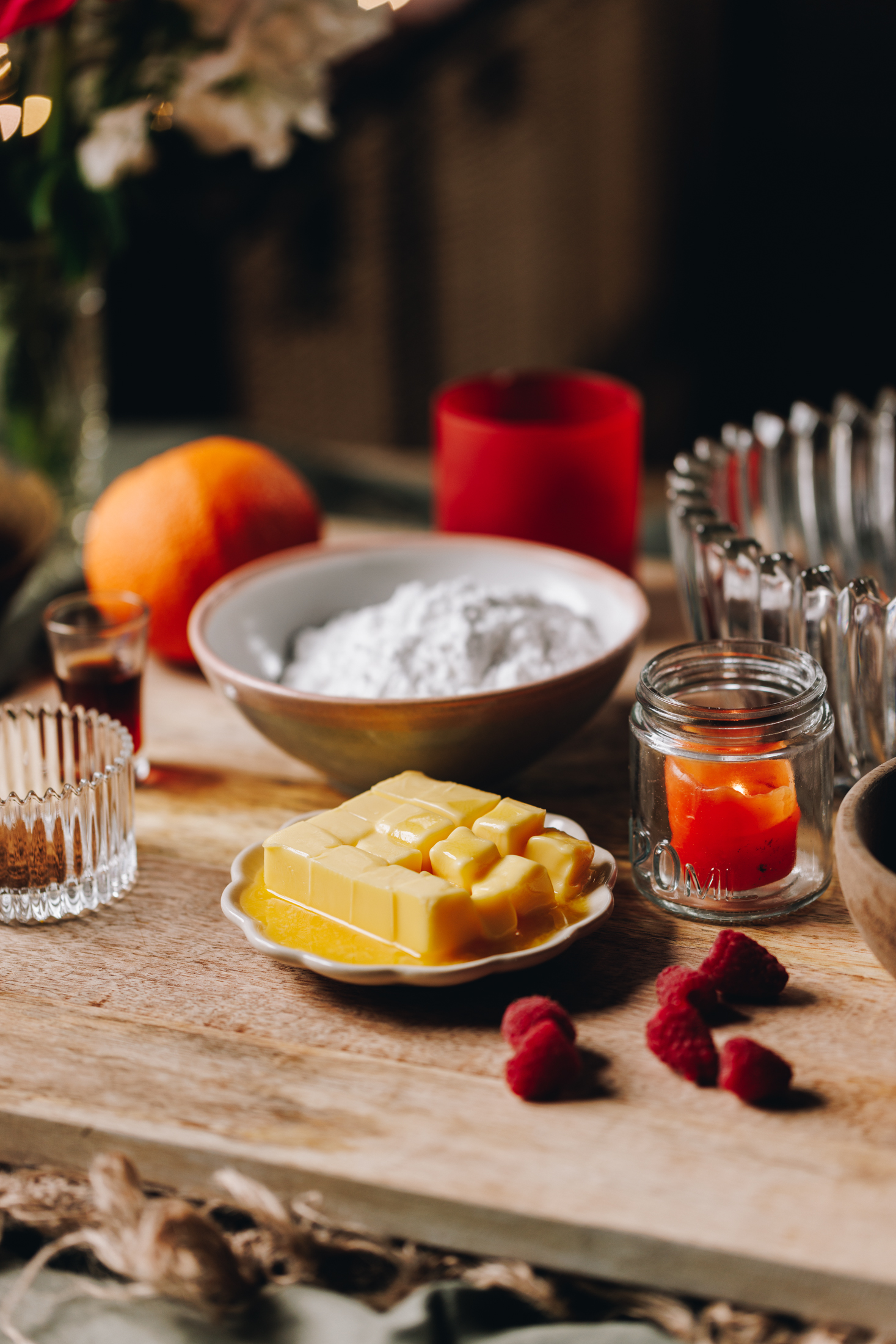 On a wooden board sits vintage bowls and small plates that have buttercream ingredients on it. There is oranges, eggs, spices and icing sugar in view with a sprinkling of fresh raspberries.