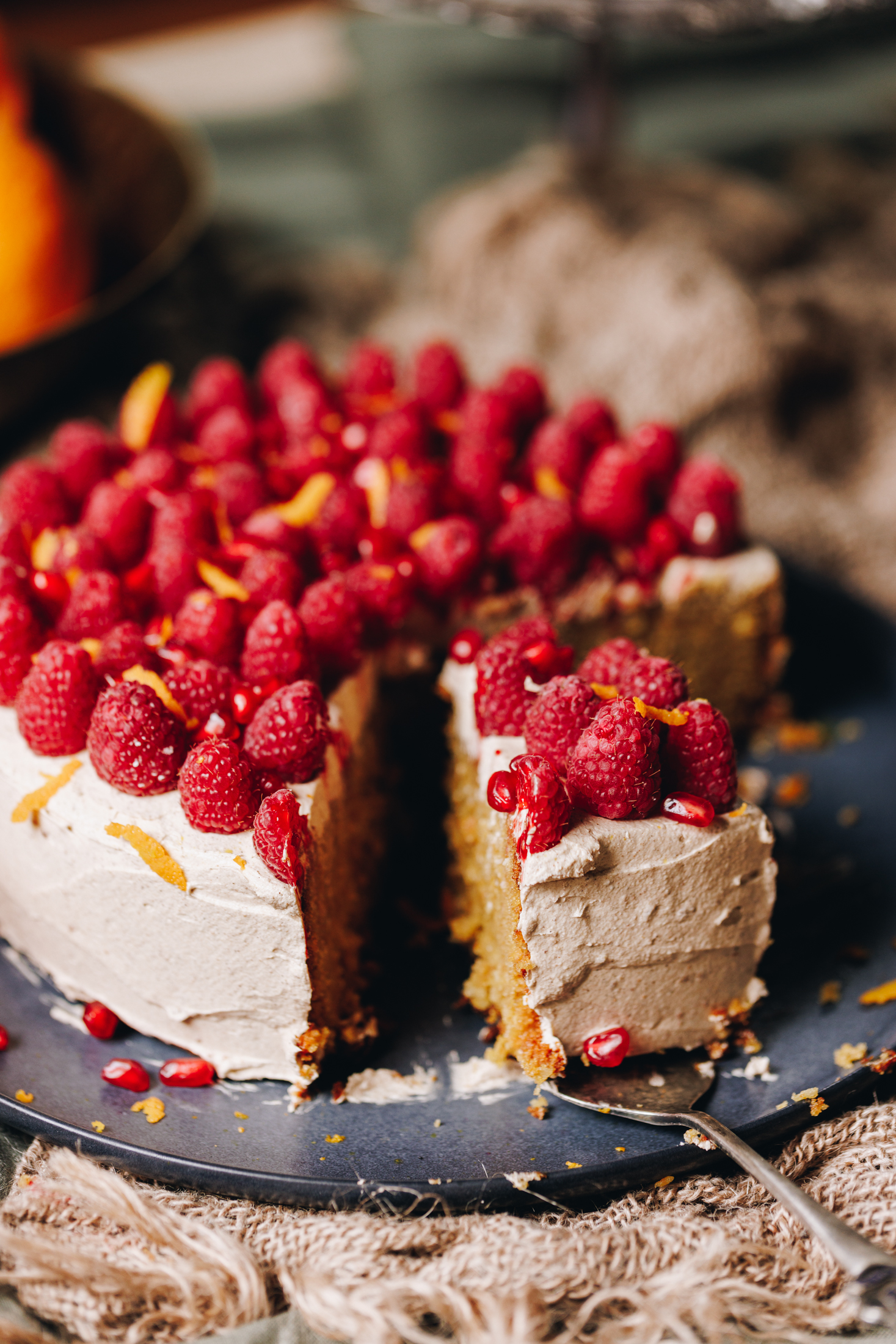 A blue plate sits on a wooden table. On the plate is a whole orange cake that is decorated in simple cinnamon buttercream with fresh raspberries covering the top of the cake with grated orange zest. The cake has been sliced open and a piece of cake is sitting on a cake server.