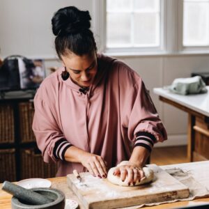Naomi Toilalo is kneading dough on a wooden board that is on a wooden table.