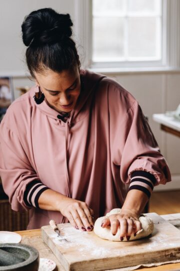 Naomi Toilalo is kneading dough on a wooden board that is on a wooden table.