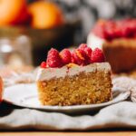 A slice of whole orange cake sits on a small white plate. It has a thick layer of simple cinnamon buttercream and fresh raspberries on top.