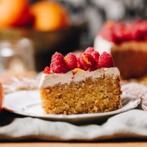 A slice of whole orange cake sits on a small white plate. It has a thick layer of simple cinnamon buttercream and fresh raspberries on top.