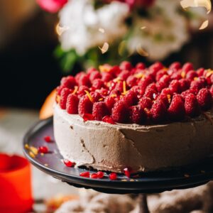 A whole orange almond cake is on a metal cake stand with a blue plate on it, the cake is decorated with cinnamon buttercream and has raspberries all over it with grated orange peel.