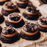Freshly baked Buckey cookies are on brown baking paper. They have a chocolate cookie, peanut butter truffle, chocolate on top with a chocolate decoration on each one.