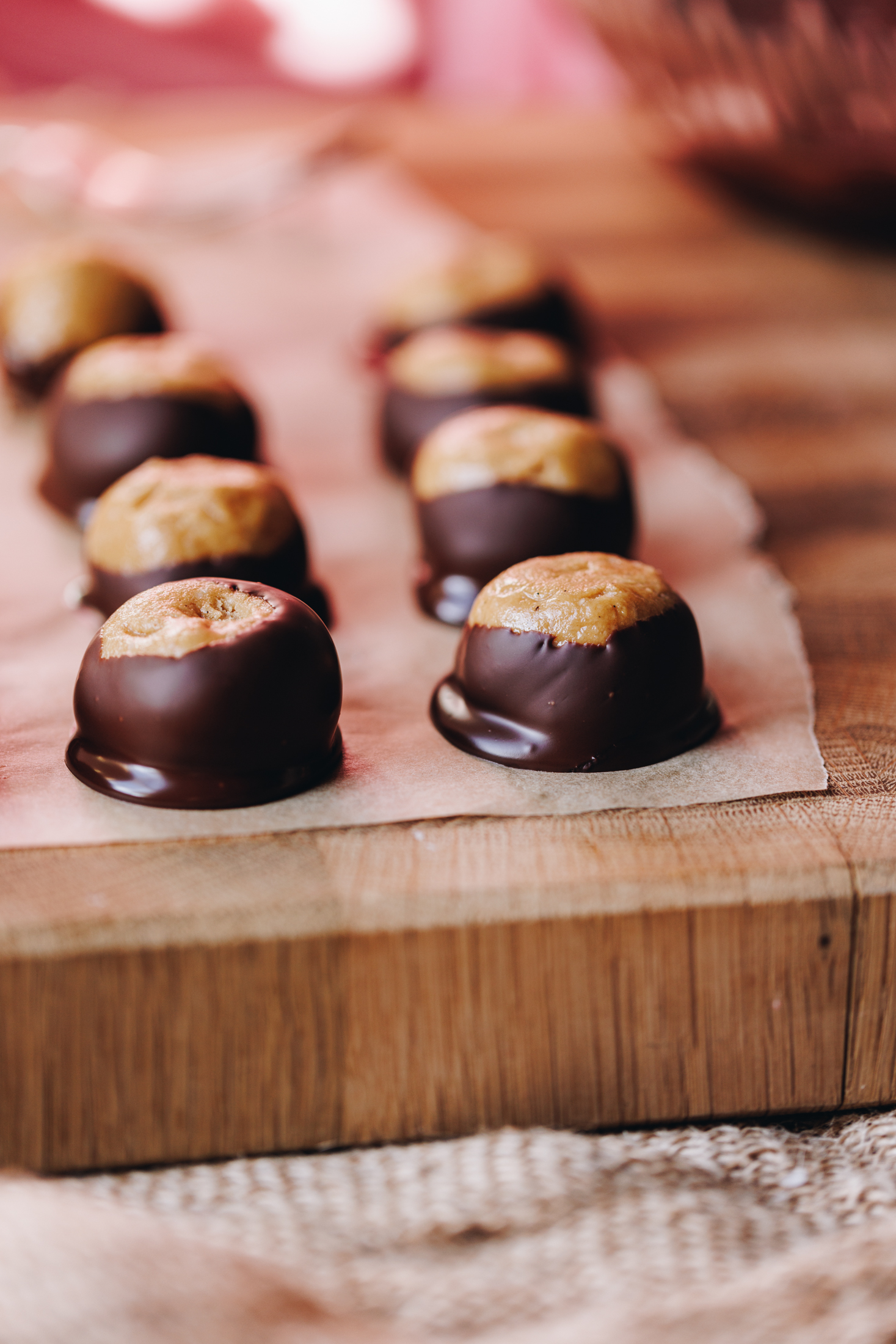 A wooden board lined with brown baking paper sits on a cloth. On the baking paper is freshly decorated peanut butter buckeye truffles. They truffles are round and they have been dipped in chocolate with the top part of the truffle exposed. 