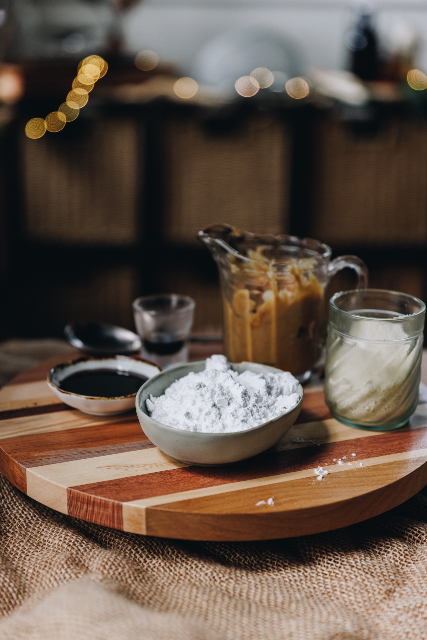 On a wooden board sits different vintage bowls and glasses holding the ingredients for the truffles. There is icing sugar, peanut butter, vanilla paste and almond meal in view. Blurred fairy lights are in the background. 