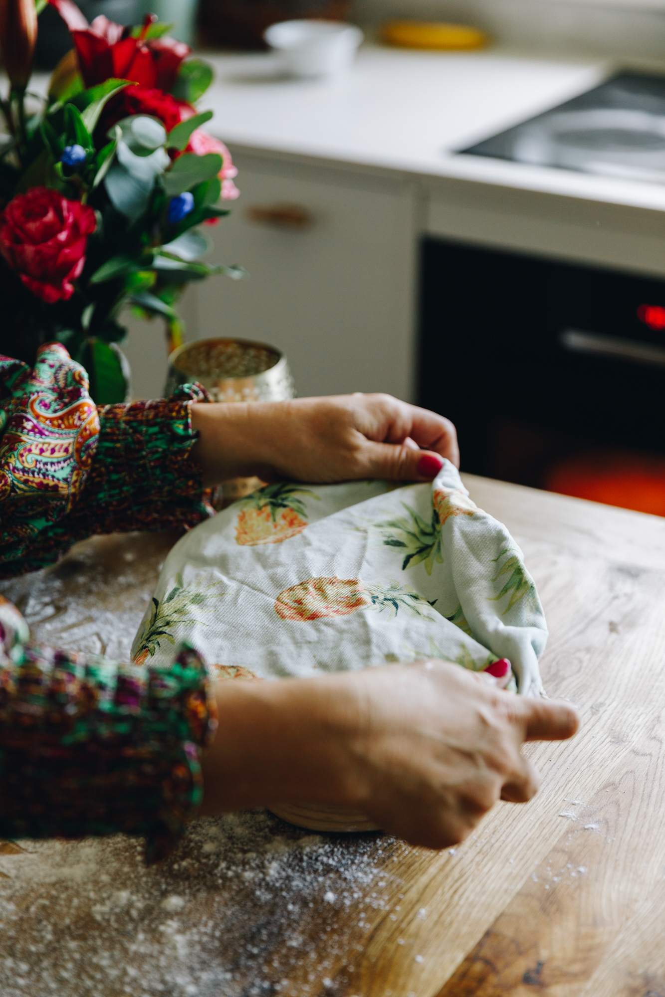 A dough is in a sourdough banetton, it sits on a wooden, floured table with flowers in the background. A bowl cover is covering the bannetton.