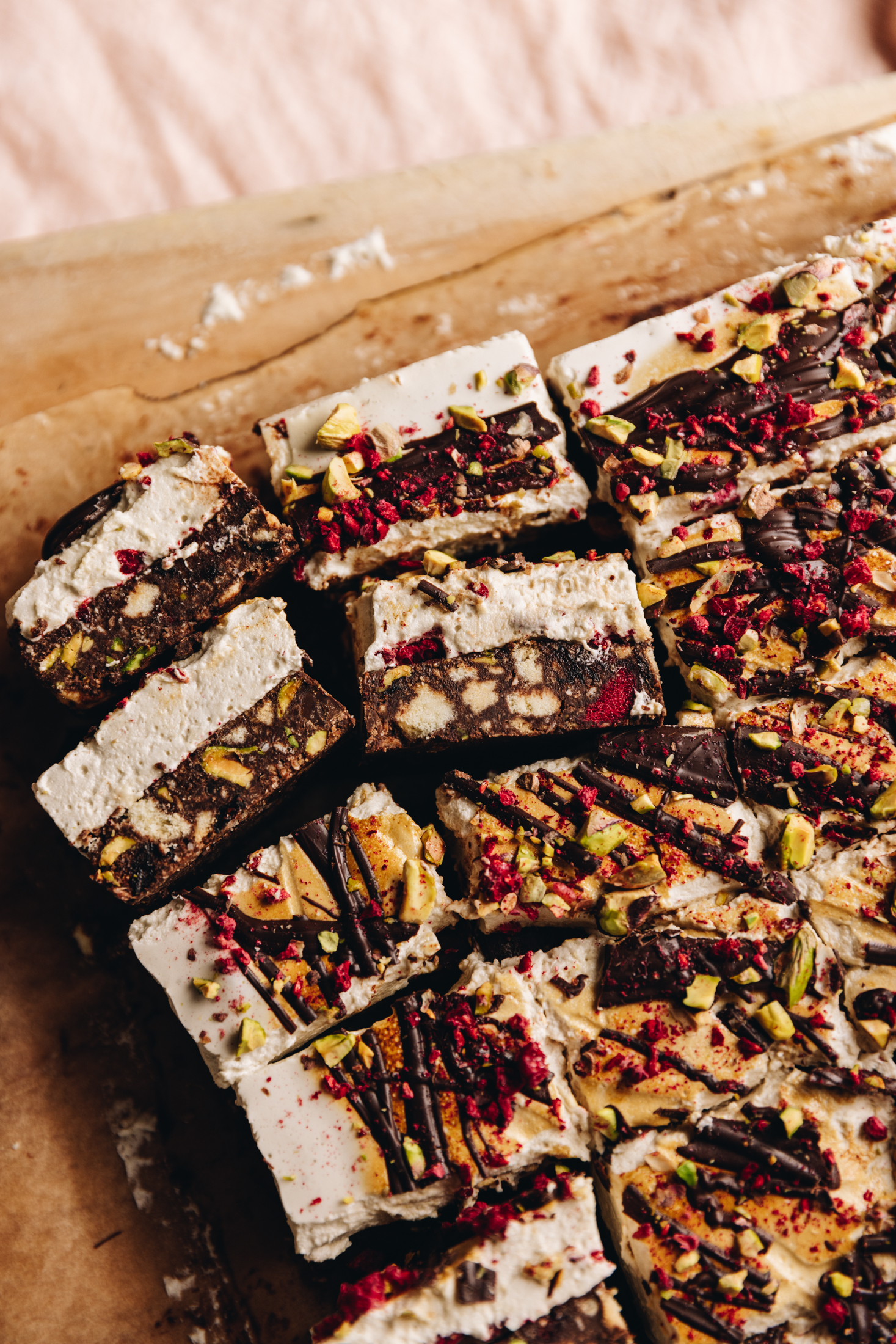 A flat lay shot shows a no-bake marshmallow hedgehog slice that has been cut in to pieces. The slice has got a marshmallow topping that has drizzled chocolate on top with freeze-dried plums and crushed pistachios. Three pieces are flipped on their side to reveal the hedgehog slice layer and the fluffy marshmallow layer. 