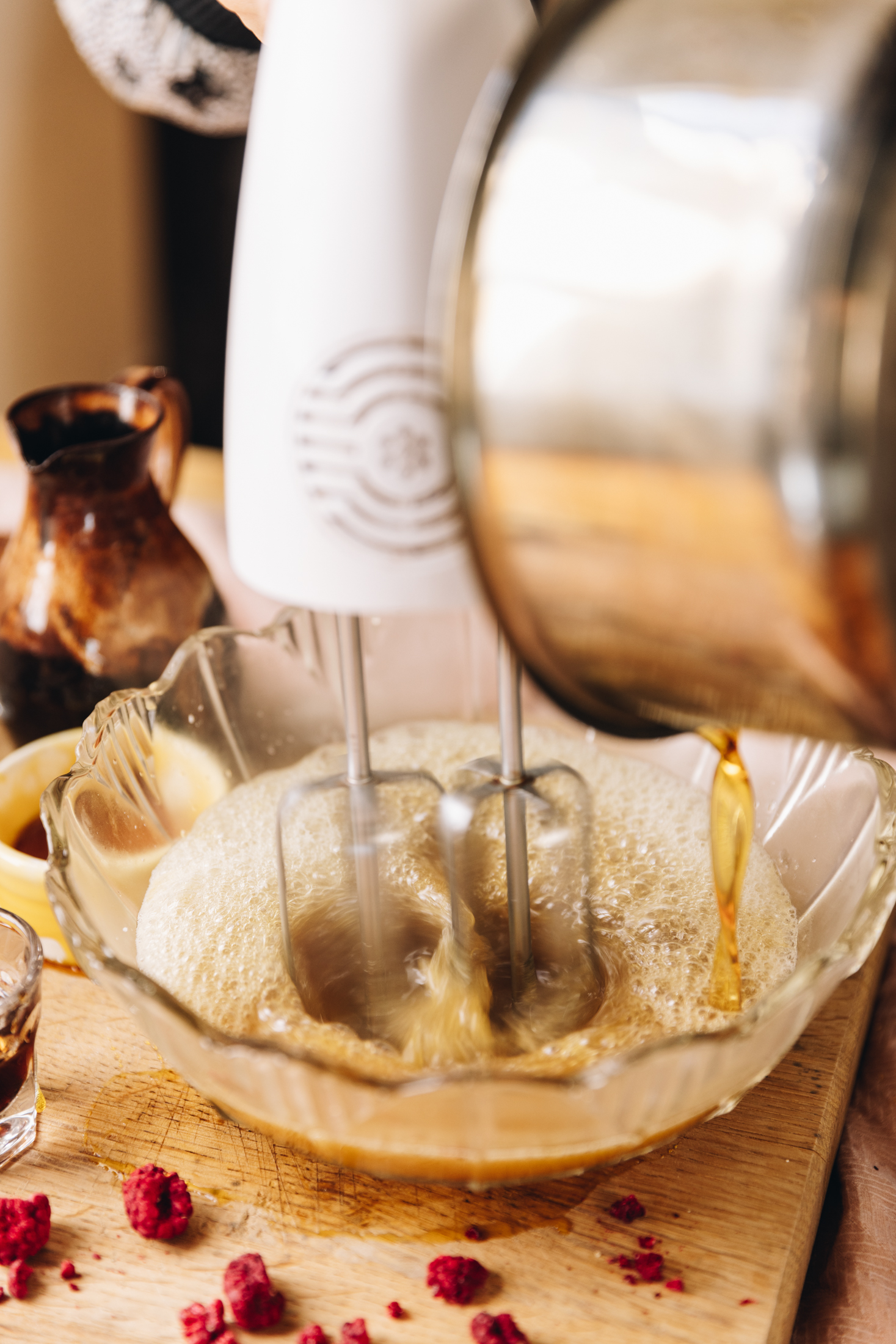 A wooden board sits on a wooden table. On the board is a glass vintage bowl that has marshmallow liquid in it. A silver pot is pouring hot sugar syrup in to the bowl as a white hand mixer whips the mixture together. Freeze-dried raspberries are also scattered on the board and a brown and black ceramic jug is in the background. 