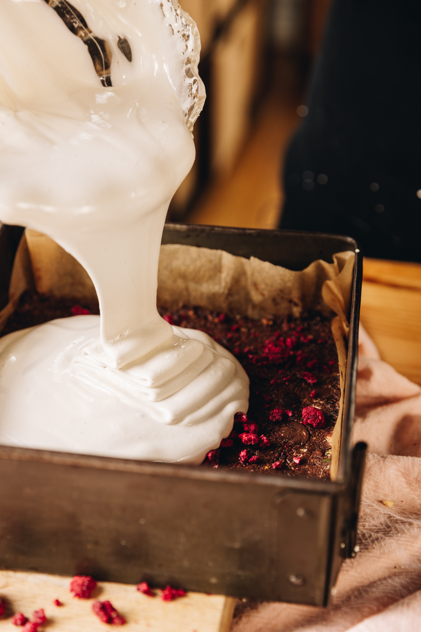 A vintage square baking tray lined with brown baking paper sits on a wooden board that is on top of a pink tablecloth, on a wooden table. In the tin is hedgehog slice that is having fresh marshmallow poured on to it from a glass bowl.