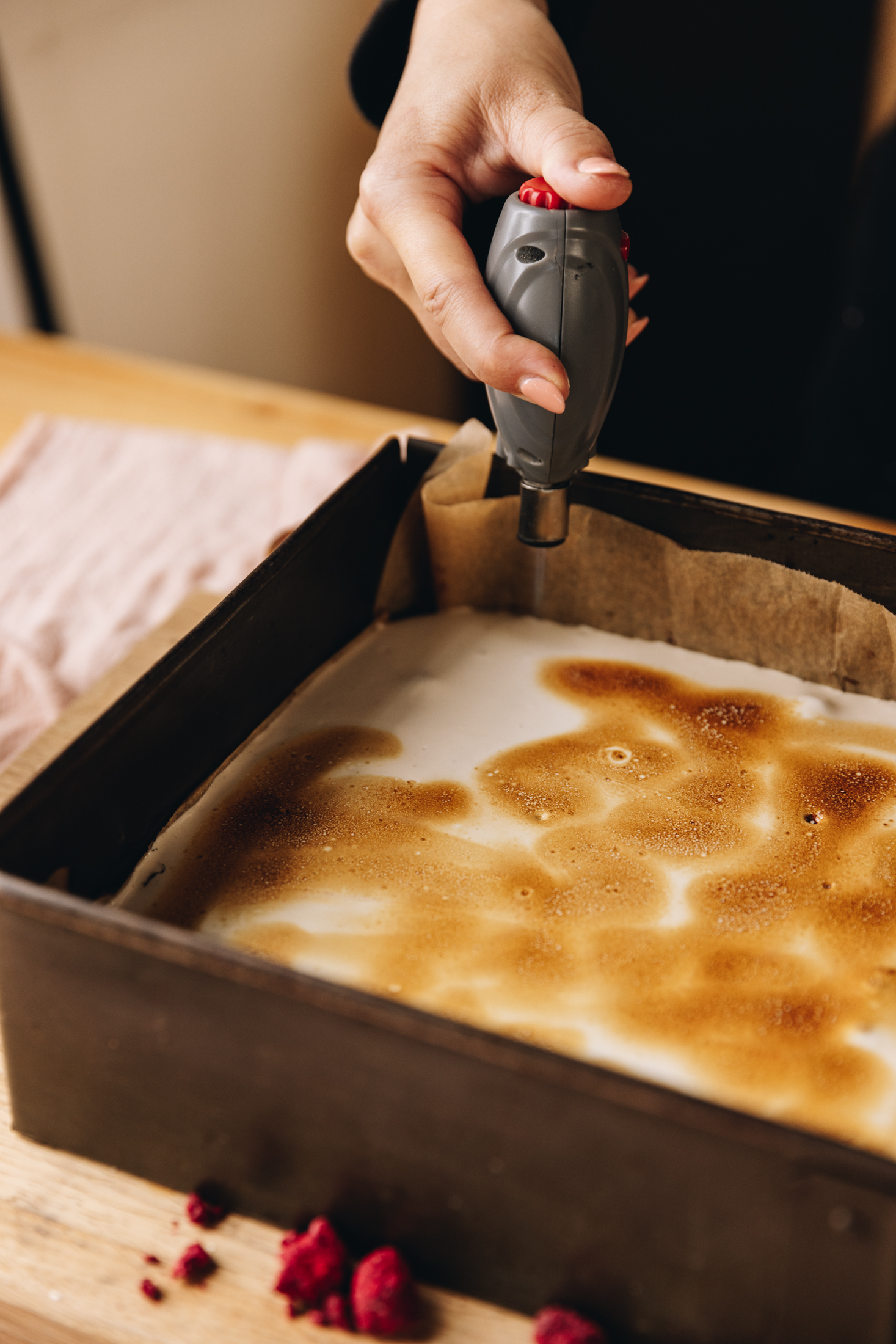 A vintage square baking tray lined with brown baking paper sits on a wooden board that is on top of a pink tablecloth, on a wooden table. In the tin is hedgehog slice that is having fresh marshmallow torched with a chef's torch. 