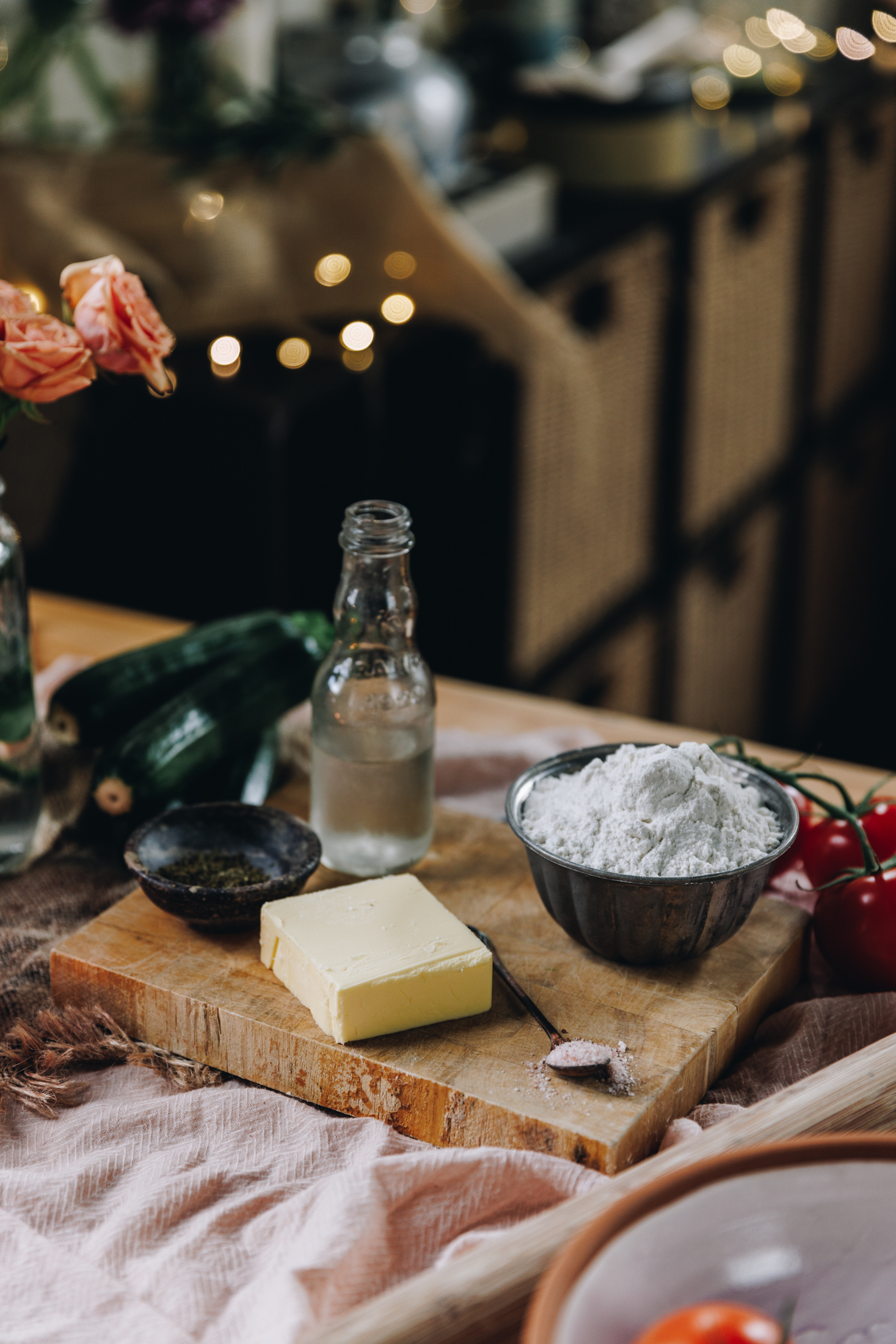 A wooden board sits on a table with a soft pink tablecloth underneath it. On the board is a square of butter, a teasoiin of salt, a dark, super small bowl with dried herbs in it, a small bow with flour in it and a glass jar with water. Behind it is a glass jar with peach flowers in it. 