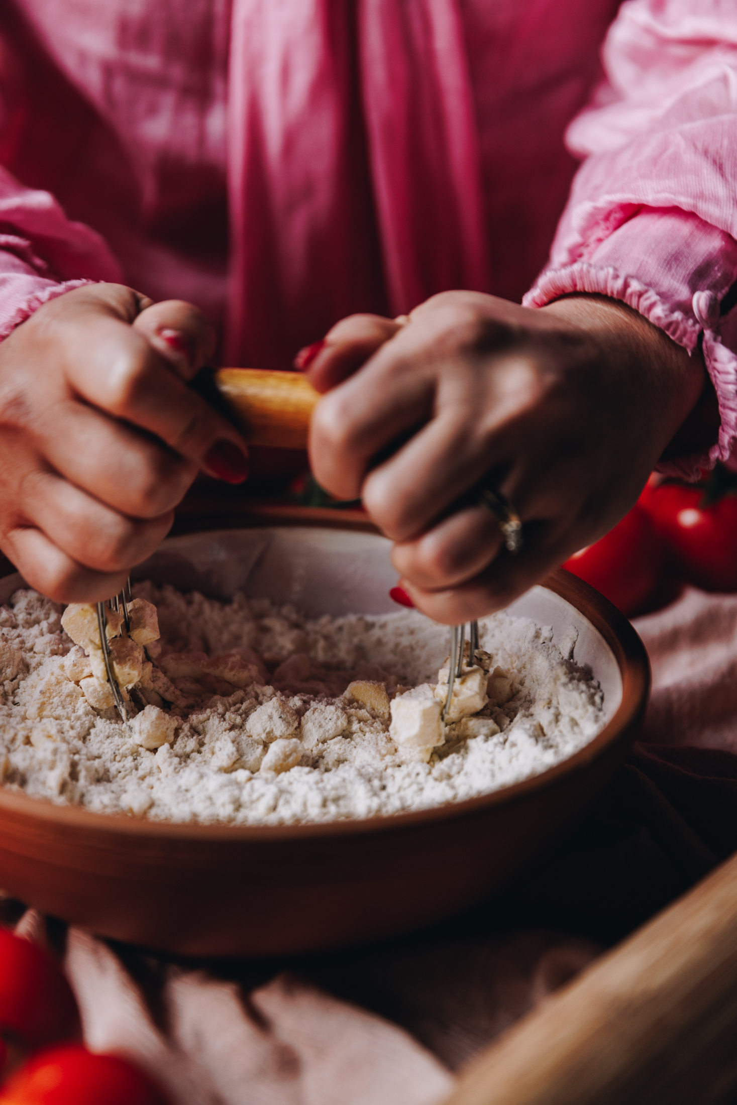 A ceramic bowl sits on a table on top of a soft pink tablecloth. In the bowl is flour and chopped butter that is being cut together with a pastry cutter. Naomi is holding with both of her hands. Fresh tomatoes can be seen in the corners of the frame and a wooden rolling pin. 