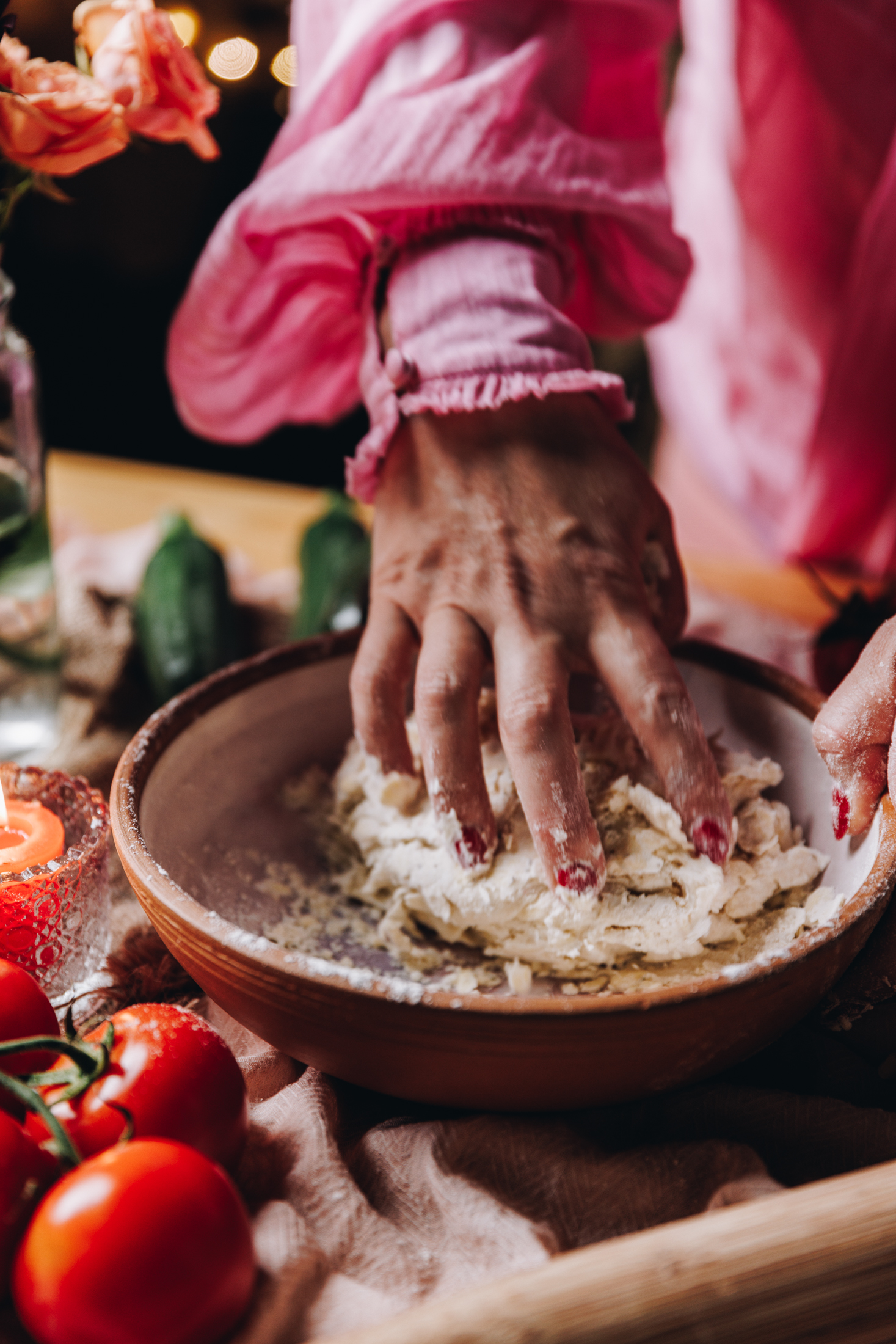 A ceramic bowl sits on a table on top of a soft pink tablecloth. In the bowl is a savoury pastry that Naomi is kneading with one of her hands. Fresh tomatoes can be seen in the corners of the frame. Two courgettes are in the background and a wooden rolling pin. 
