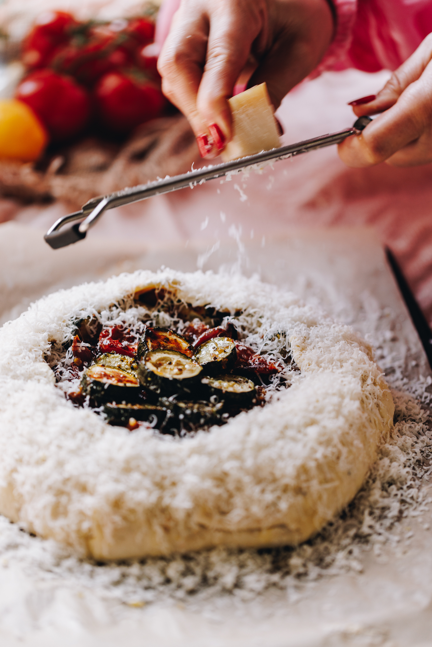 A tomato and courgette galette is wrapped in herbed shortcrust savoury pastry. Naomi is grating Parmesan cheese around the edge of the pastry with a fine grater. Tomatoes are seen in the background. 