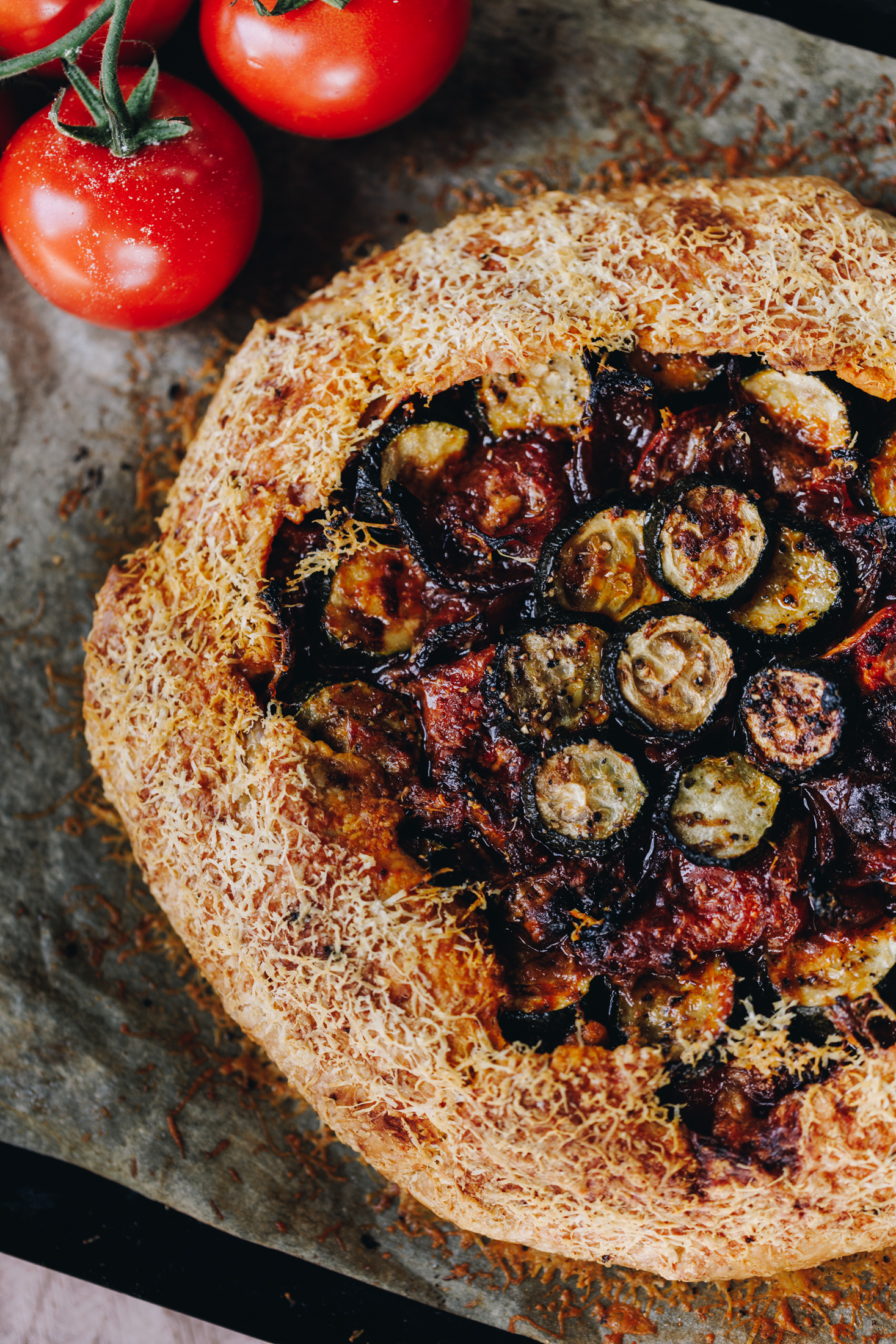 A flat lay shot shows a tomato galette that is wrapped in herbed shortcrust savoury pastry. Fresh tomatoes can be seen in the top left corner. The galette is uncut. 