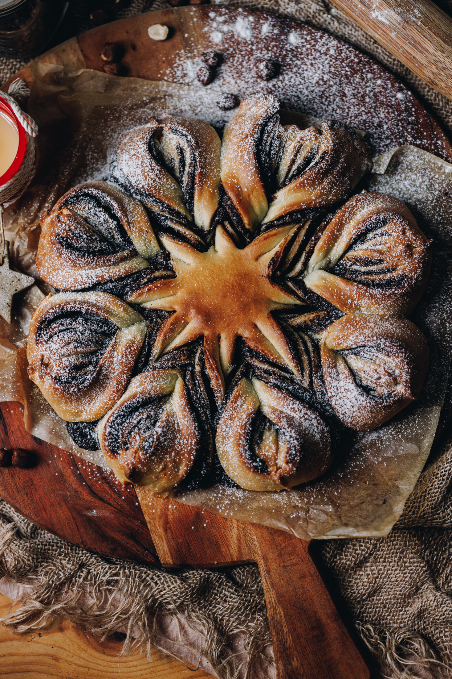 A flat lay of a freshly baked chocolate star bread is on a wooden board, dusted with icing sugar and a red candle next to it. It is on a wooden table.