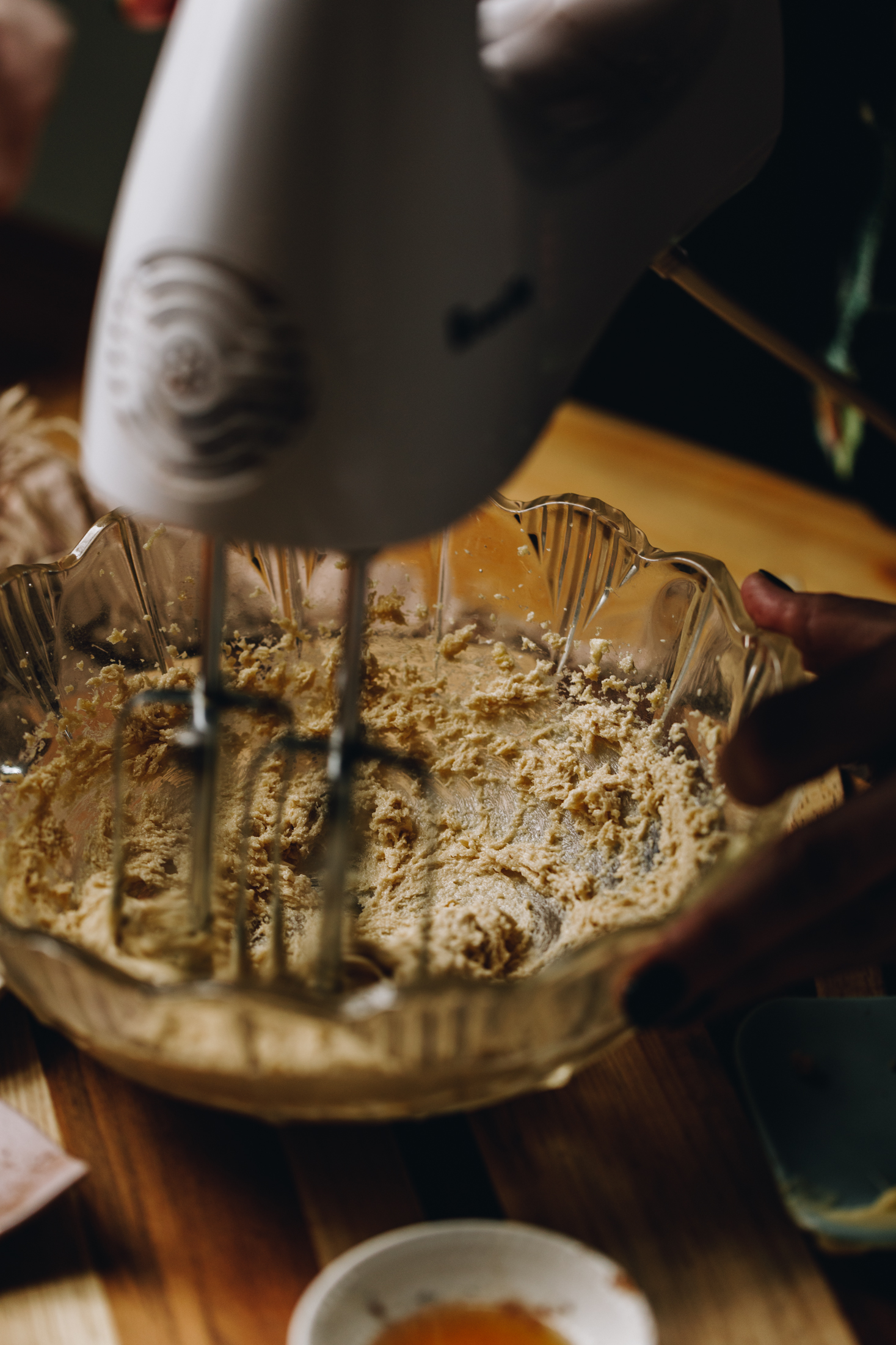 In a vintage glass bowl butter and sugar is being whipped with a white hand mixer. It is on a wooden table.