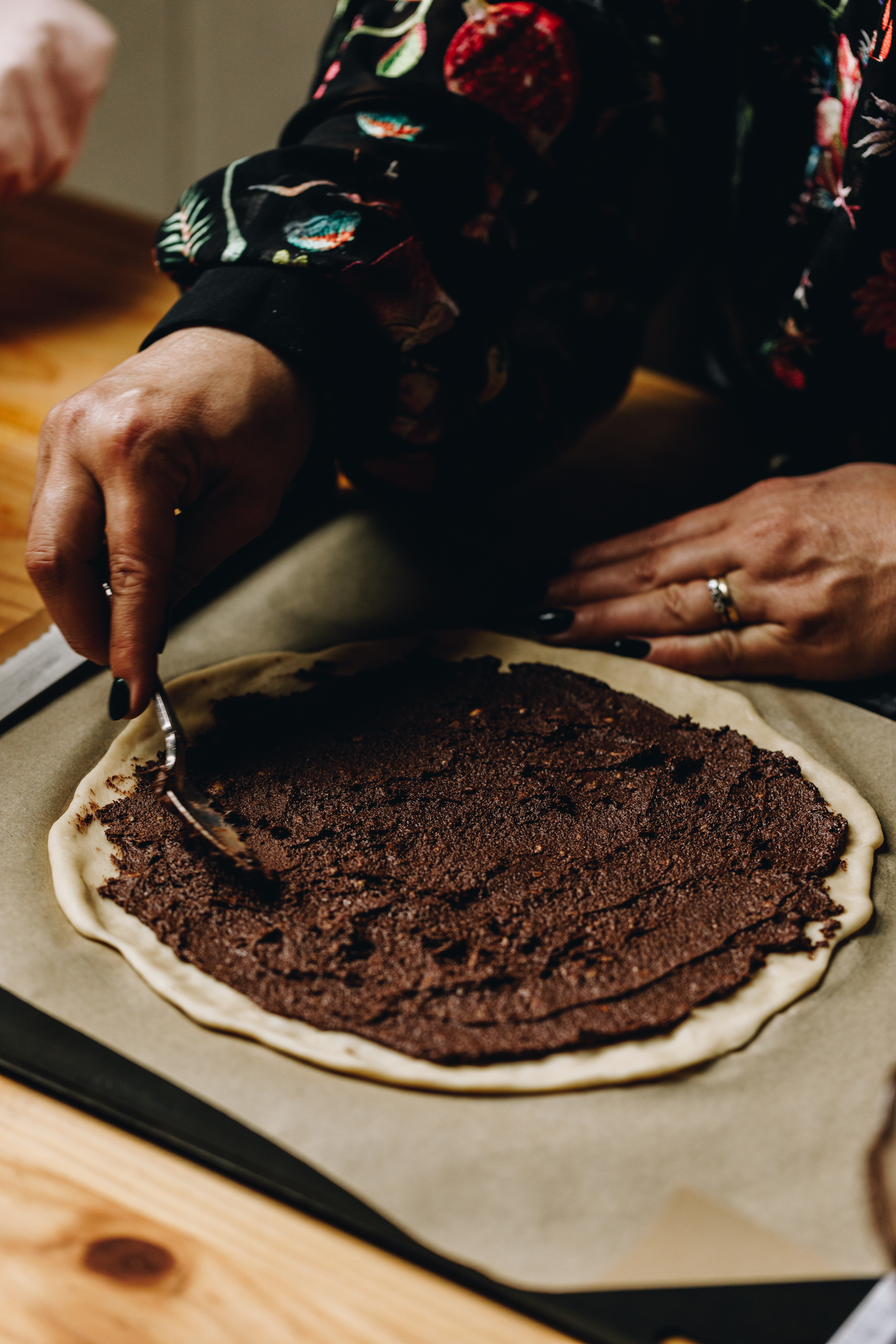 On a black tray with brown baking paper sits a large circle of dough, it is having chocolate spread spread on to it. It is on a wooden table. This is the first stage of the Chocolate Star Bread.